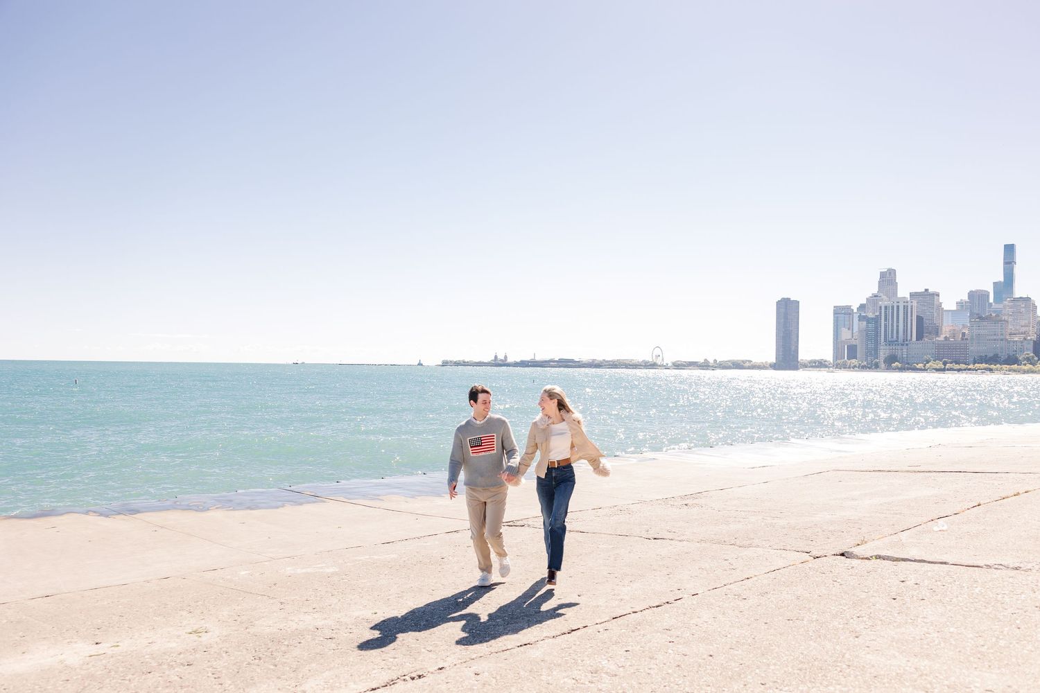 Happy couple walking hand in hand along a lakeside path with the Chicago skyline visible in the distance.