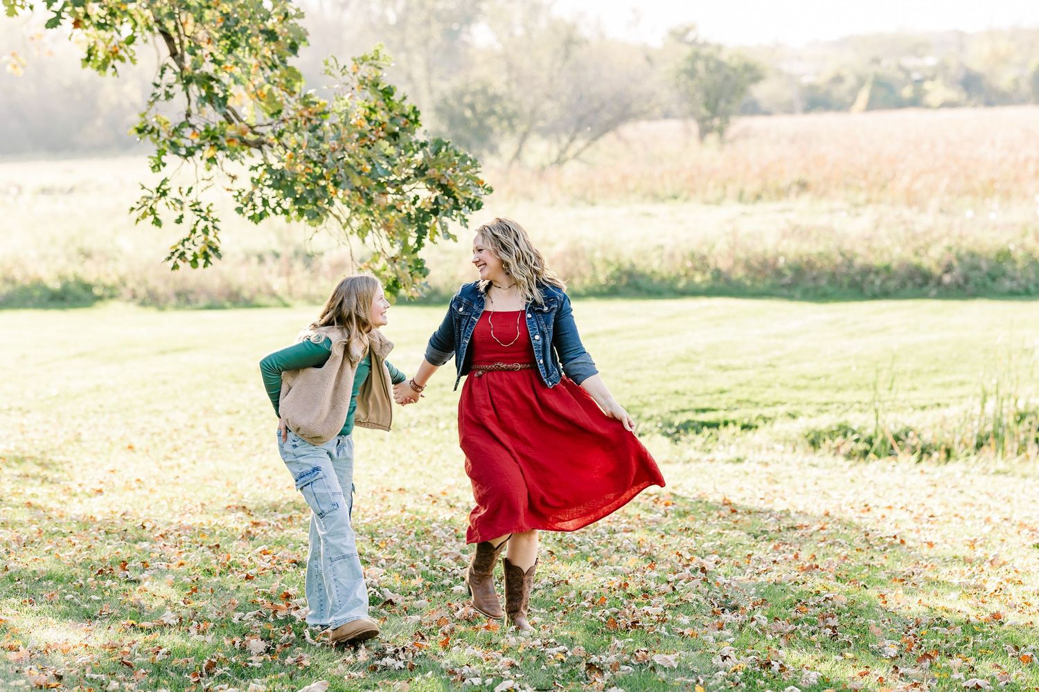 Woman in a red dress and denim jacket holds hands with a child as they walk through a sunny grassy field in autumn.