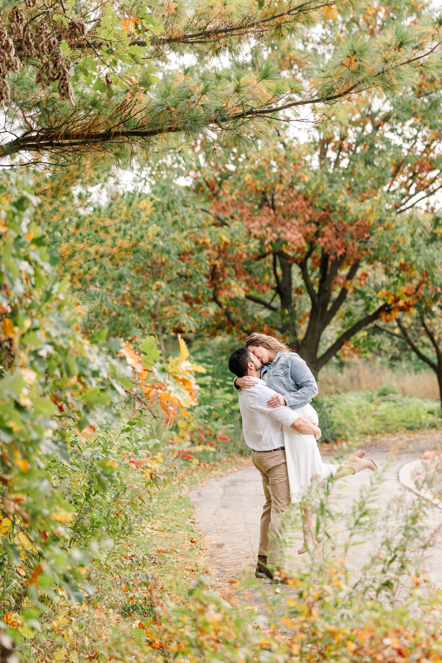 Couple embracing as he lifts her off the ground on a path surrounded by colorful autumn foliage during an engagement photo session.