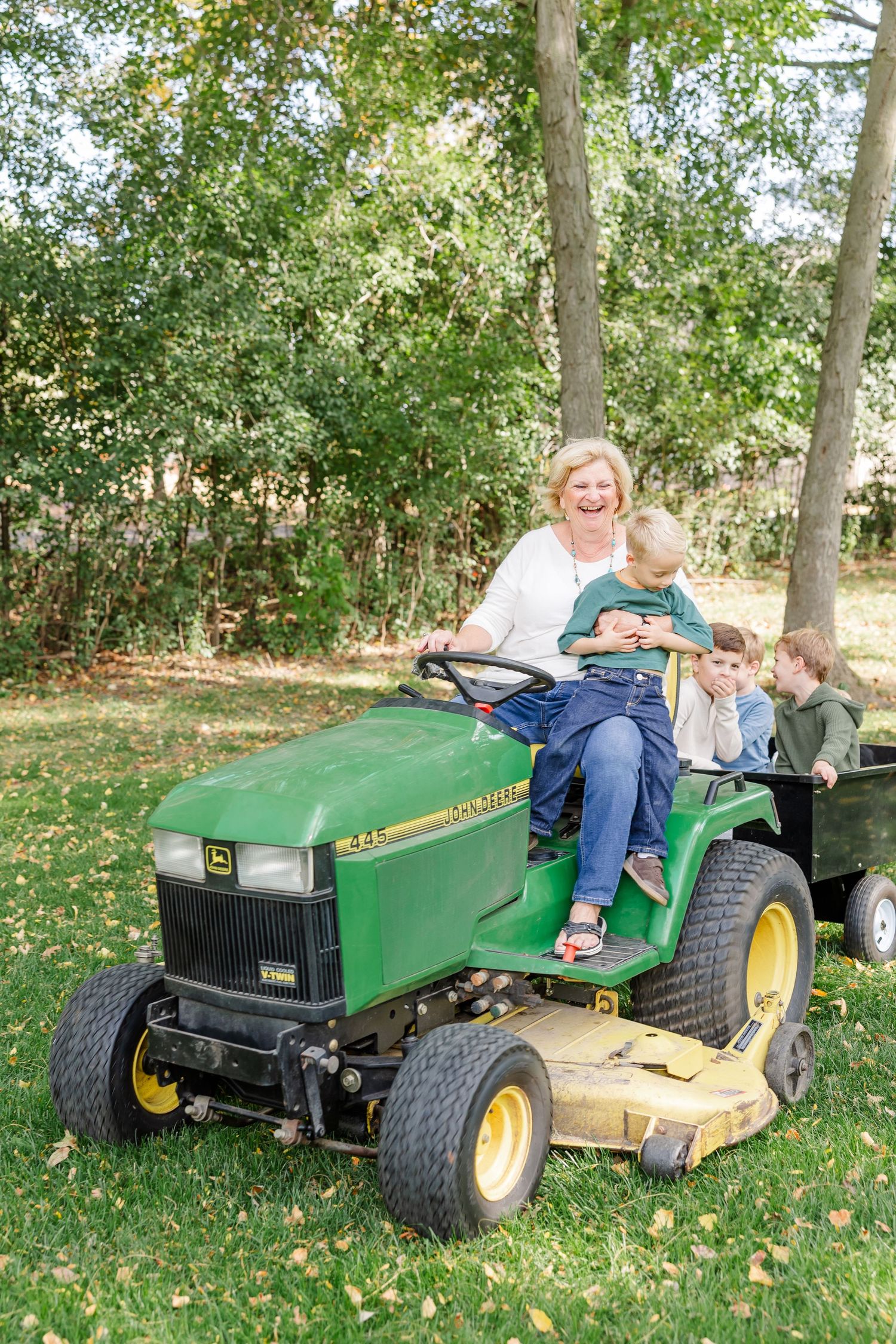 Grandmother laughing with grandchildren while riding a green John Deere tractor in a sunny backyard.