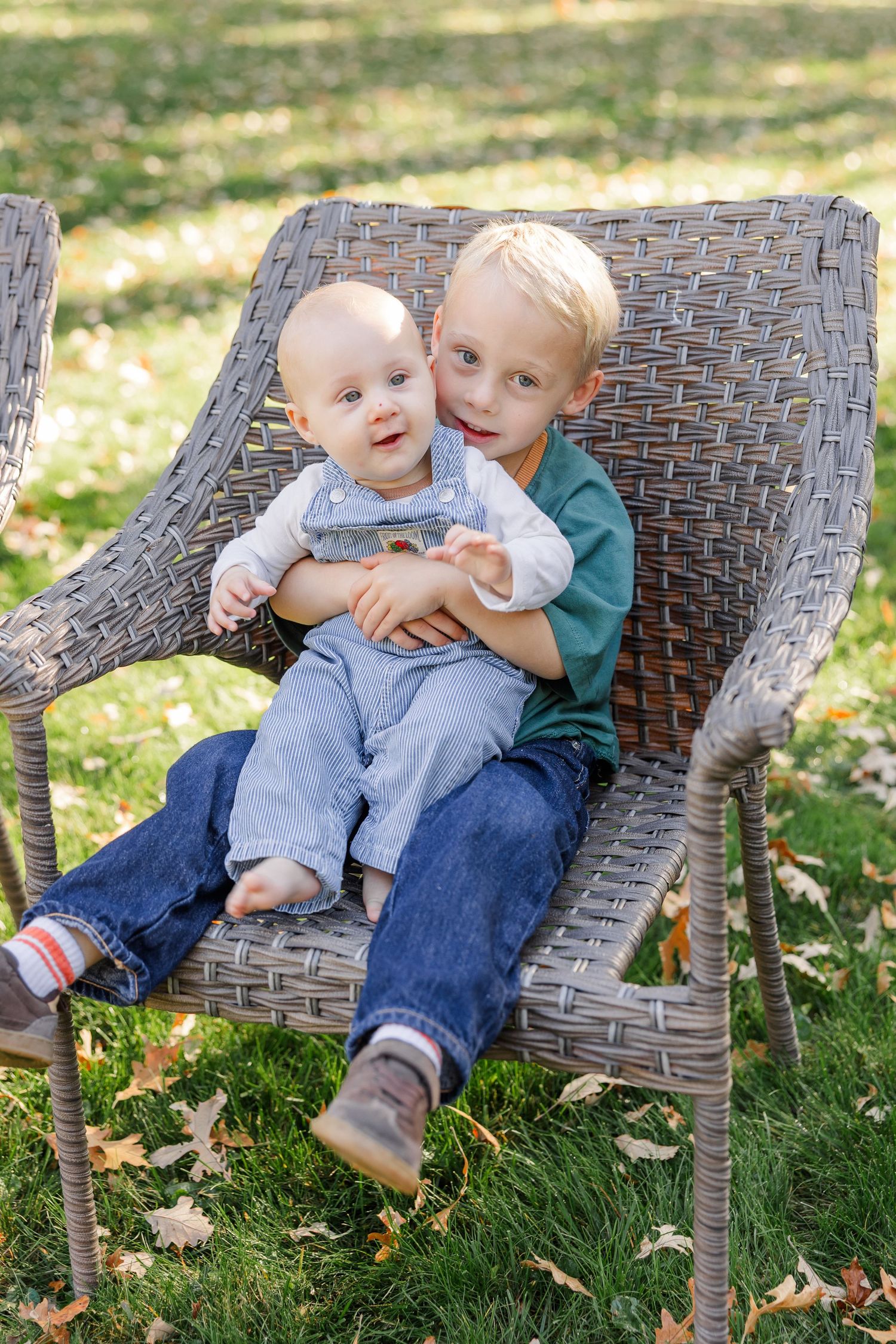Two young brothers sitting together in a wicker chair outdoors, older boy hugging baby sibling on a green lawn.