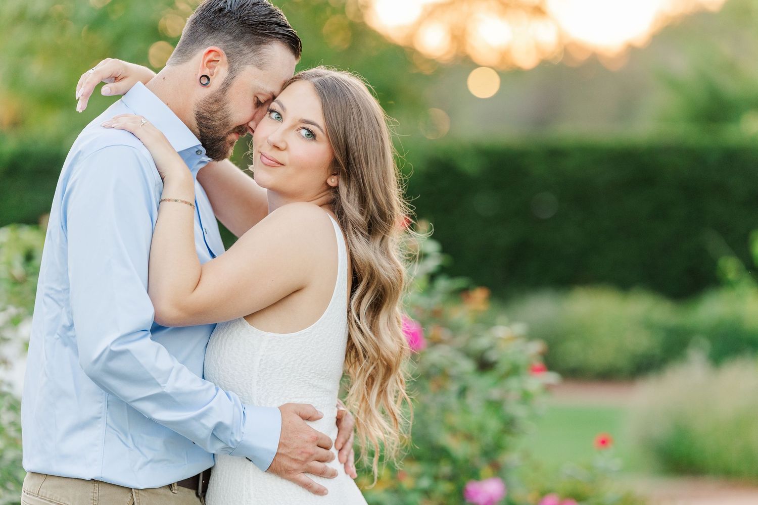 A couple embraces in a garden setting during sunset with soft golden light and blooming flowers.