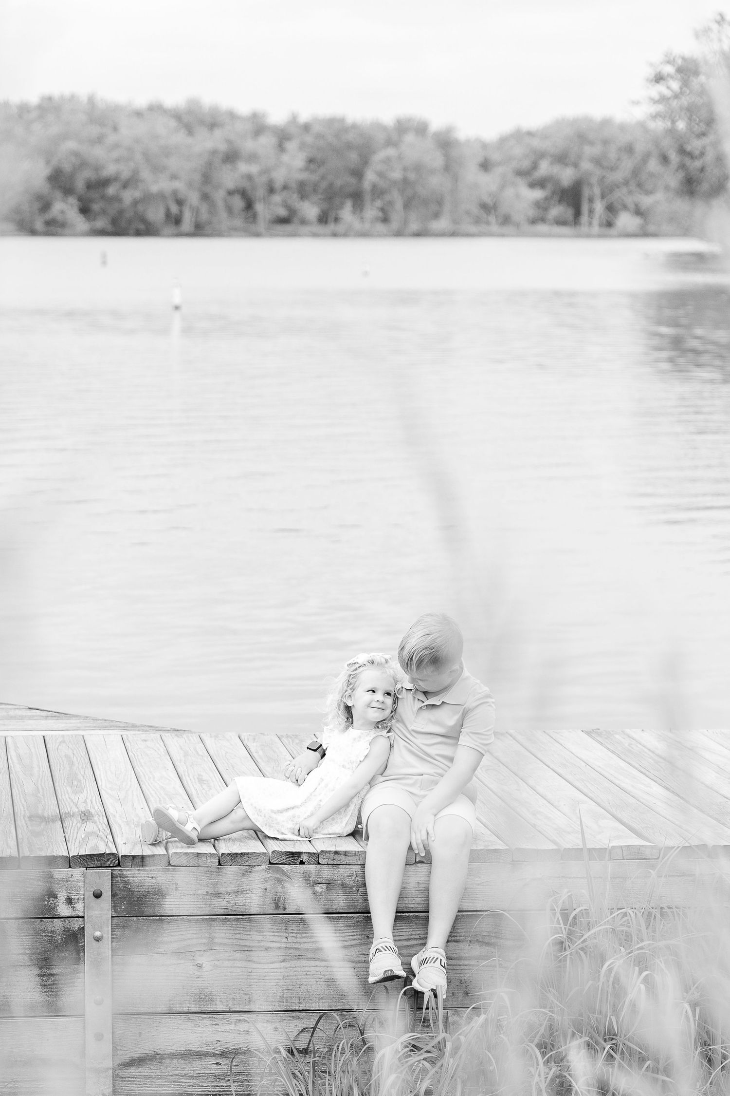 A black and white image of people sitting on a wooden dock by a peaceful lake.