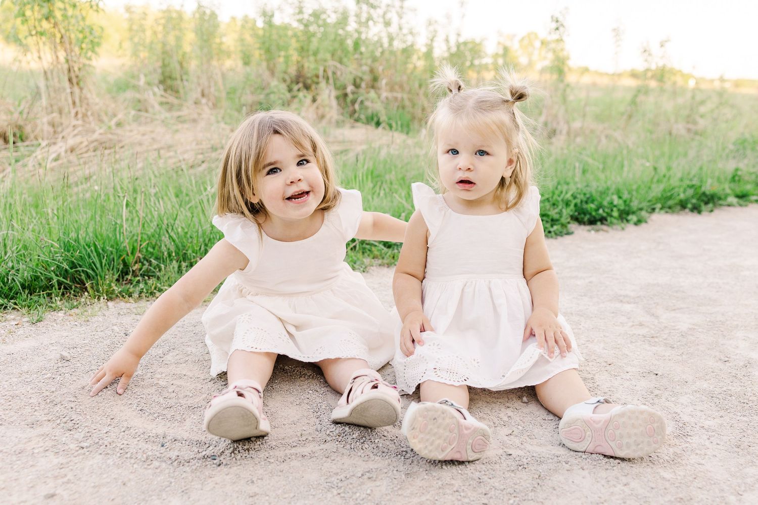 Two young children in matching light pink dresses sitting together on a dirt path.