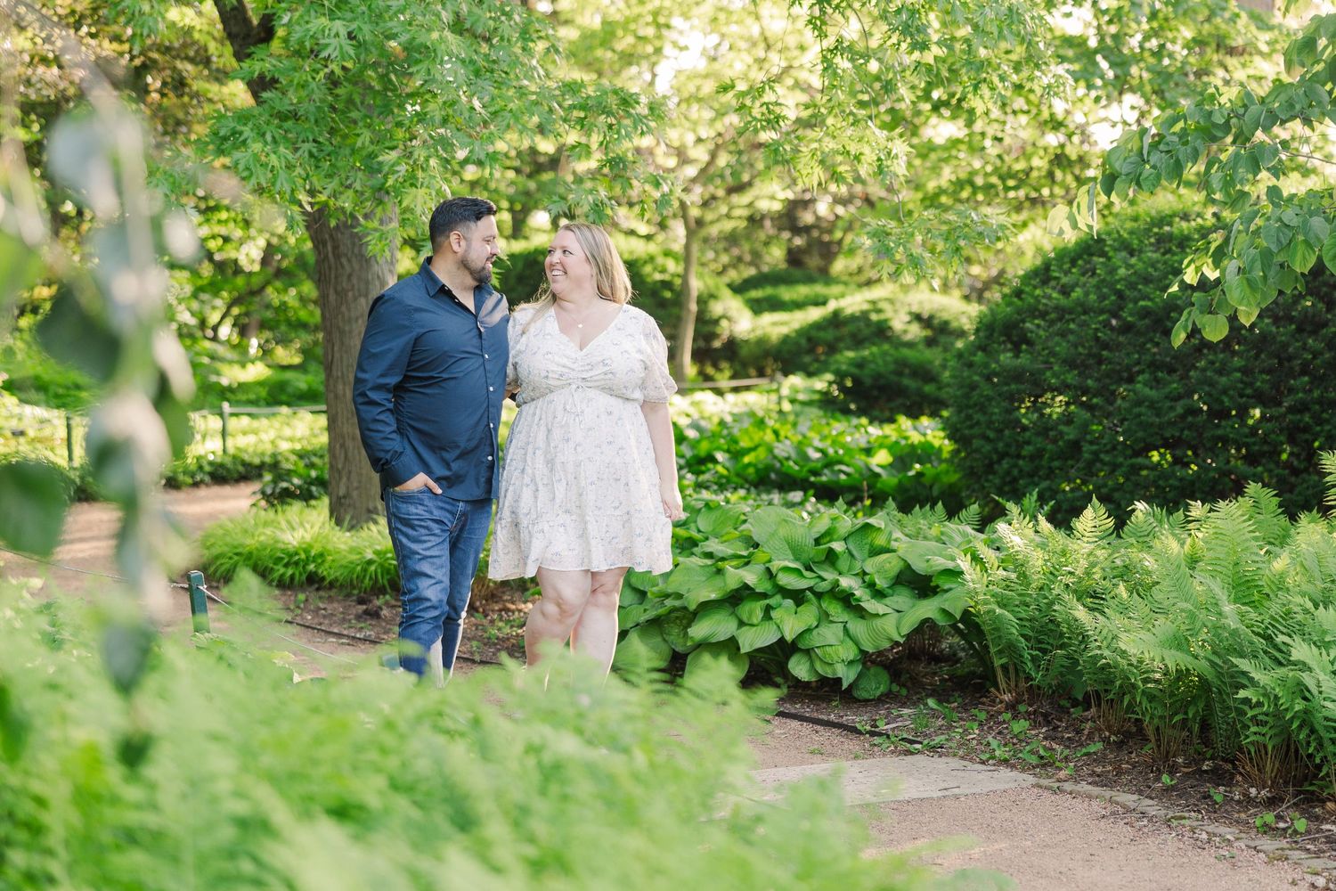 A couple strolls together along a lush green garden path during a summer engagement photo session.