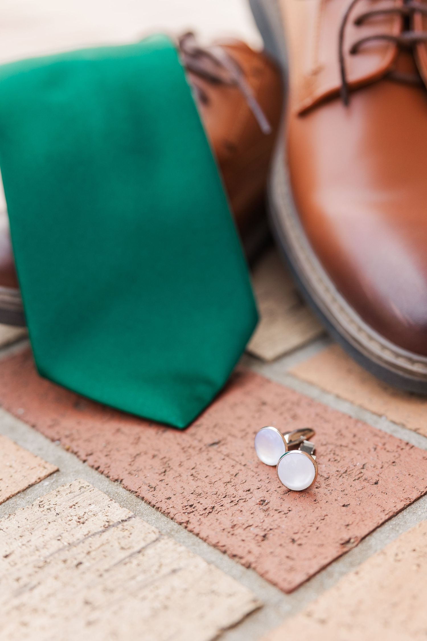 Emerald green tie with brown leather dress shoes and silver cufflinks on brick flooring.