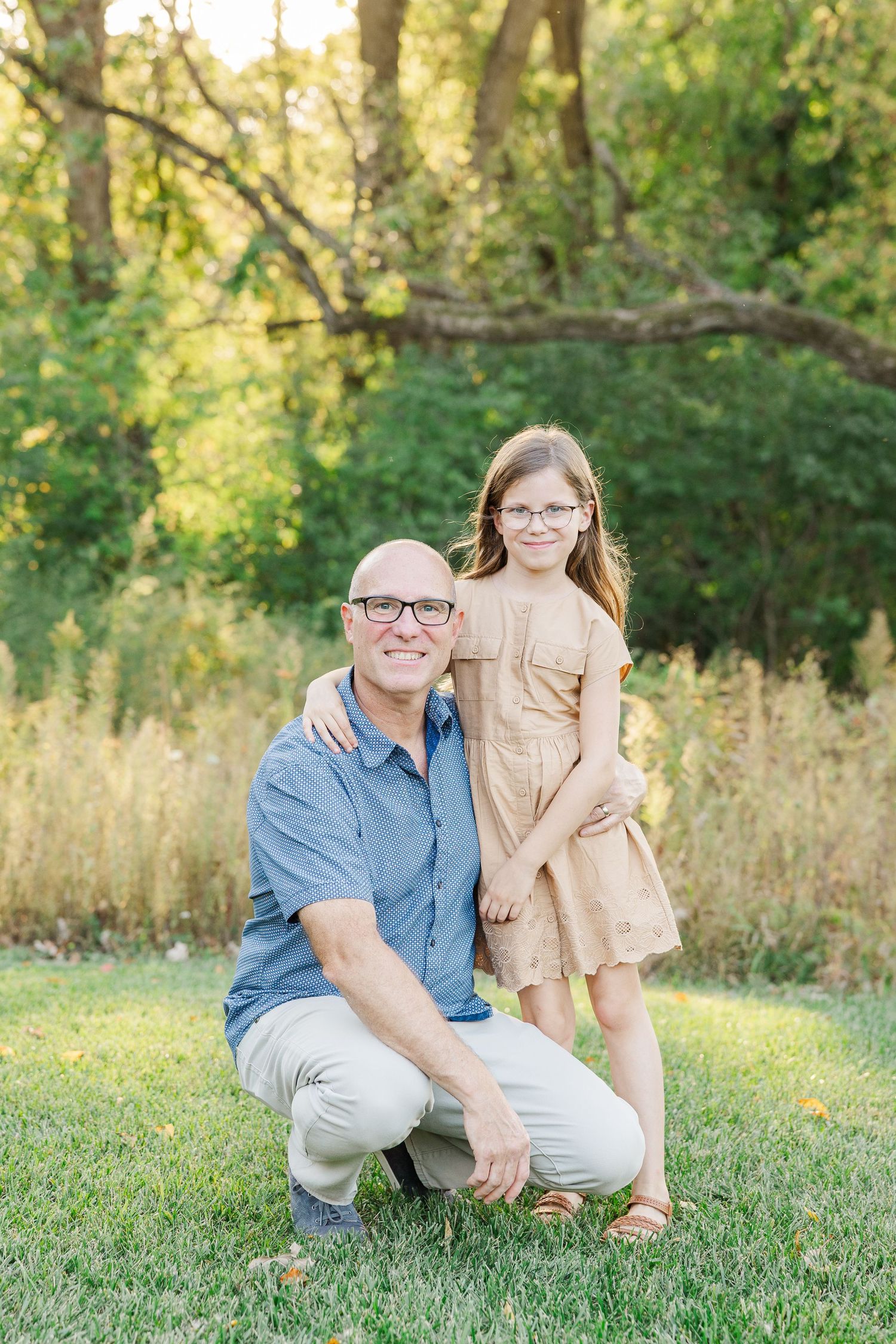 Two people sit together on lush green grass in a sunlit wooded area during a casual outdoor photo session.