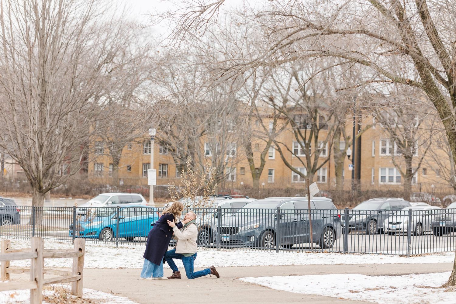 A snowy winter proposal scene with two people in silhouette against a backdrop of trees and parked cars in a lot.