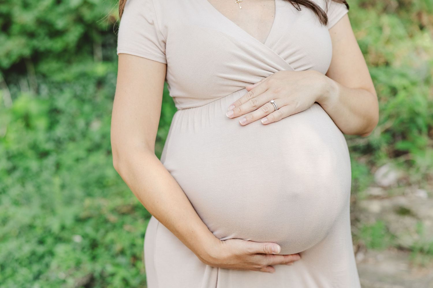 In an outdoor setting, a person in a cream-colored dress holds their pregnant belly while standing amid lush greenery.