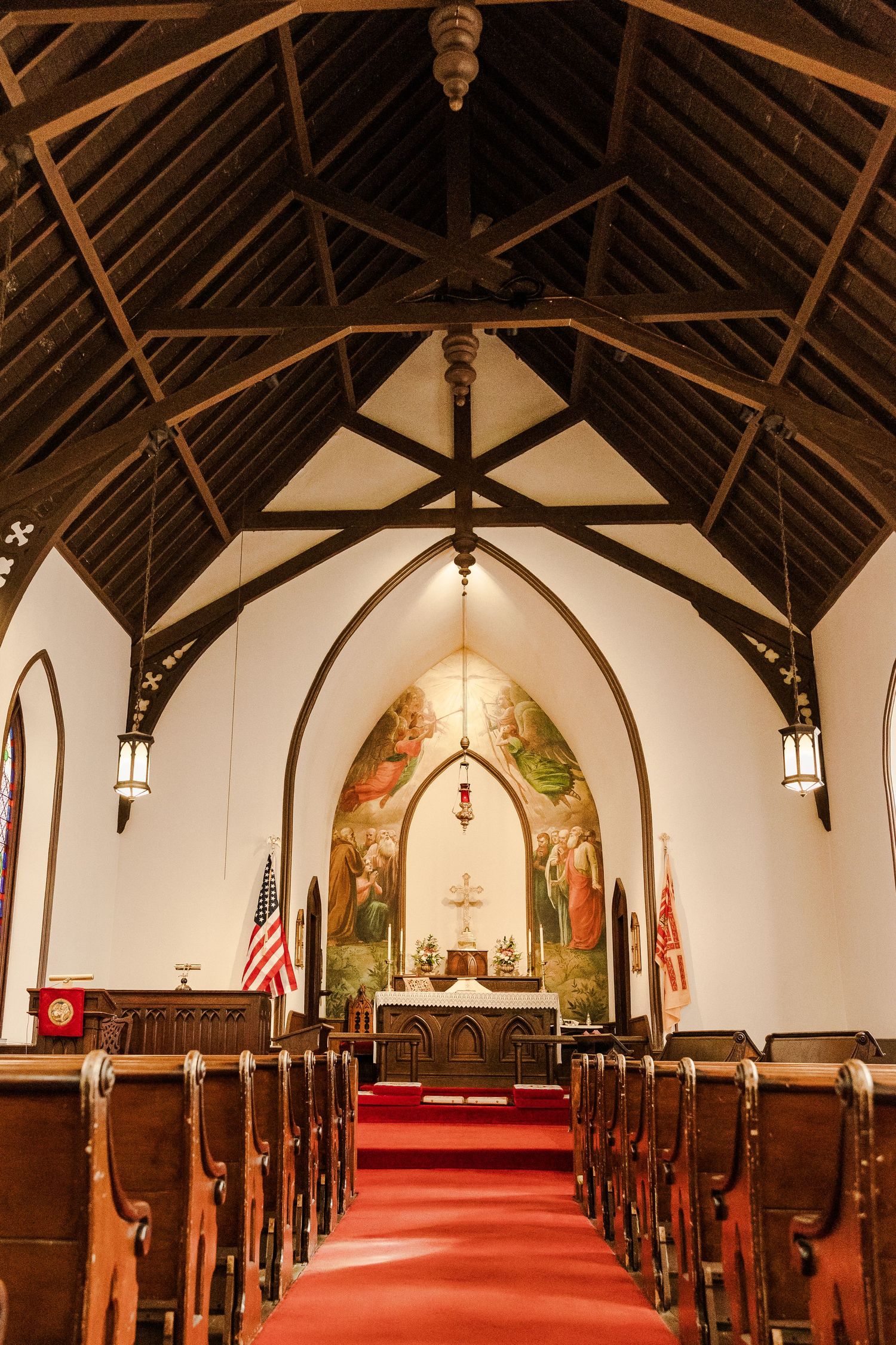 Interior of a historic church with wooden beams, red carpet, and painted altar mural in Gothic architectural style.