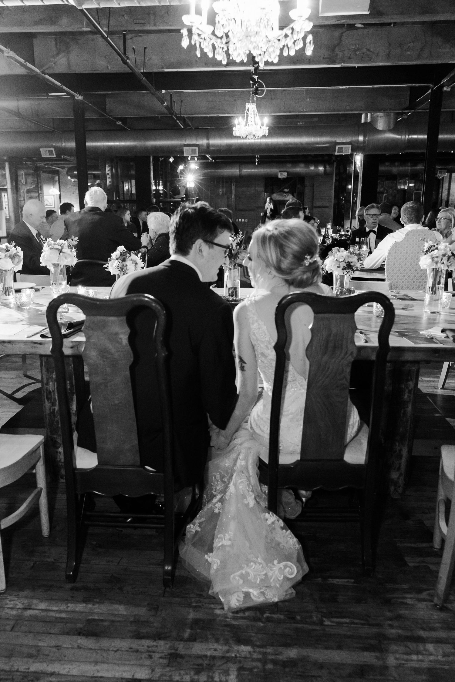 Couple seated at wedding reception under crystal chandeliers in black and white photograph.