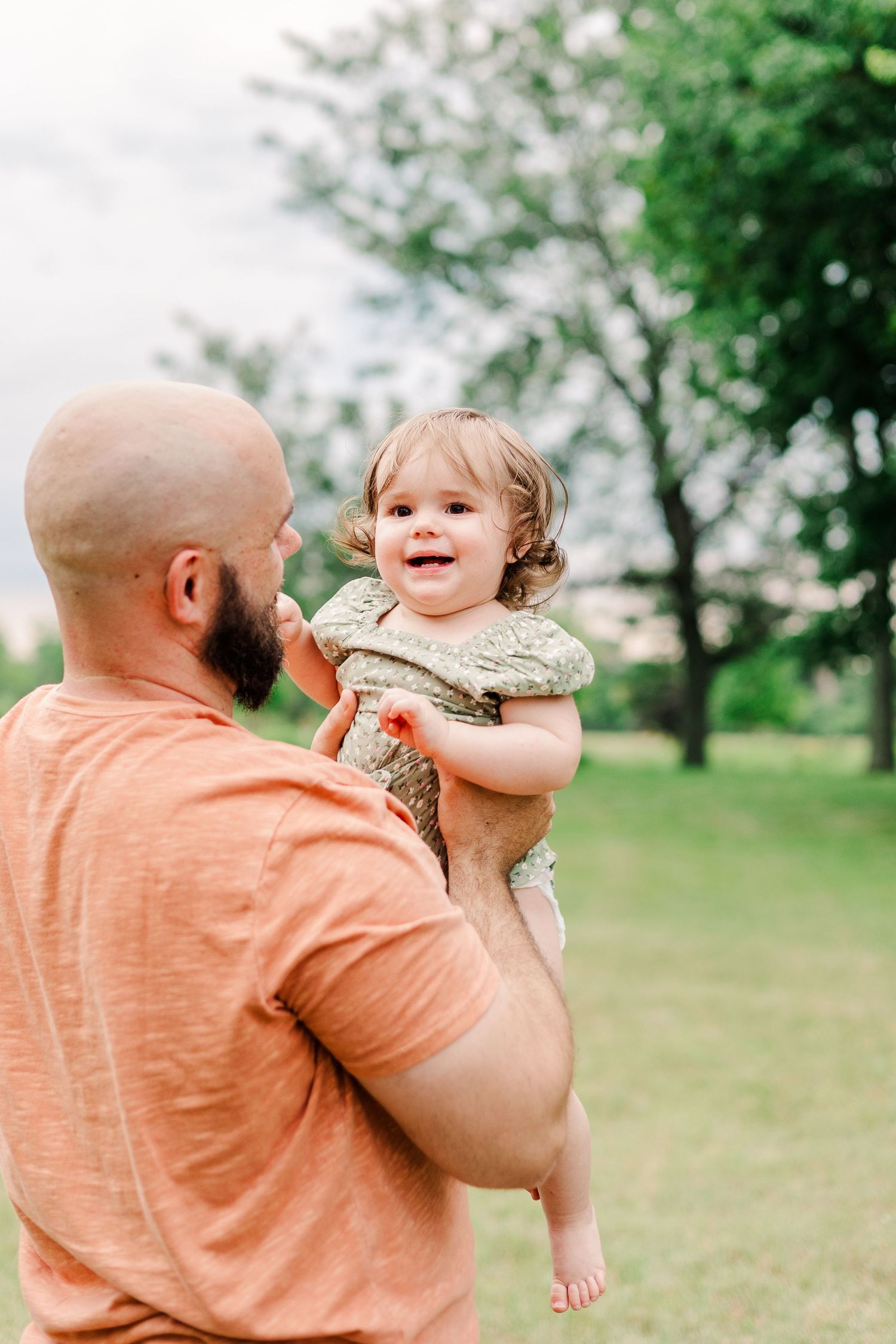 A laughing toddler in green being held up by someone in a peach shirt in a grassy outdoor setting.
