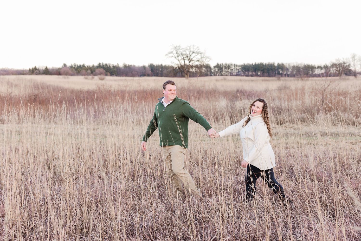 A couple holds hands while walking through a golden field of tall grass at sunset.