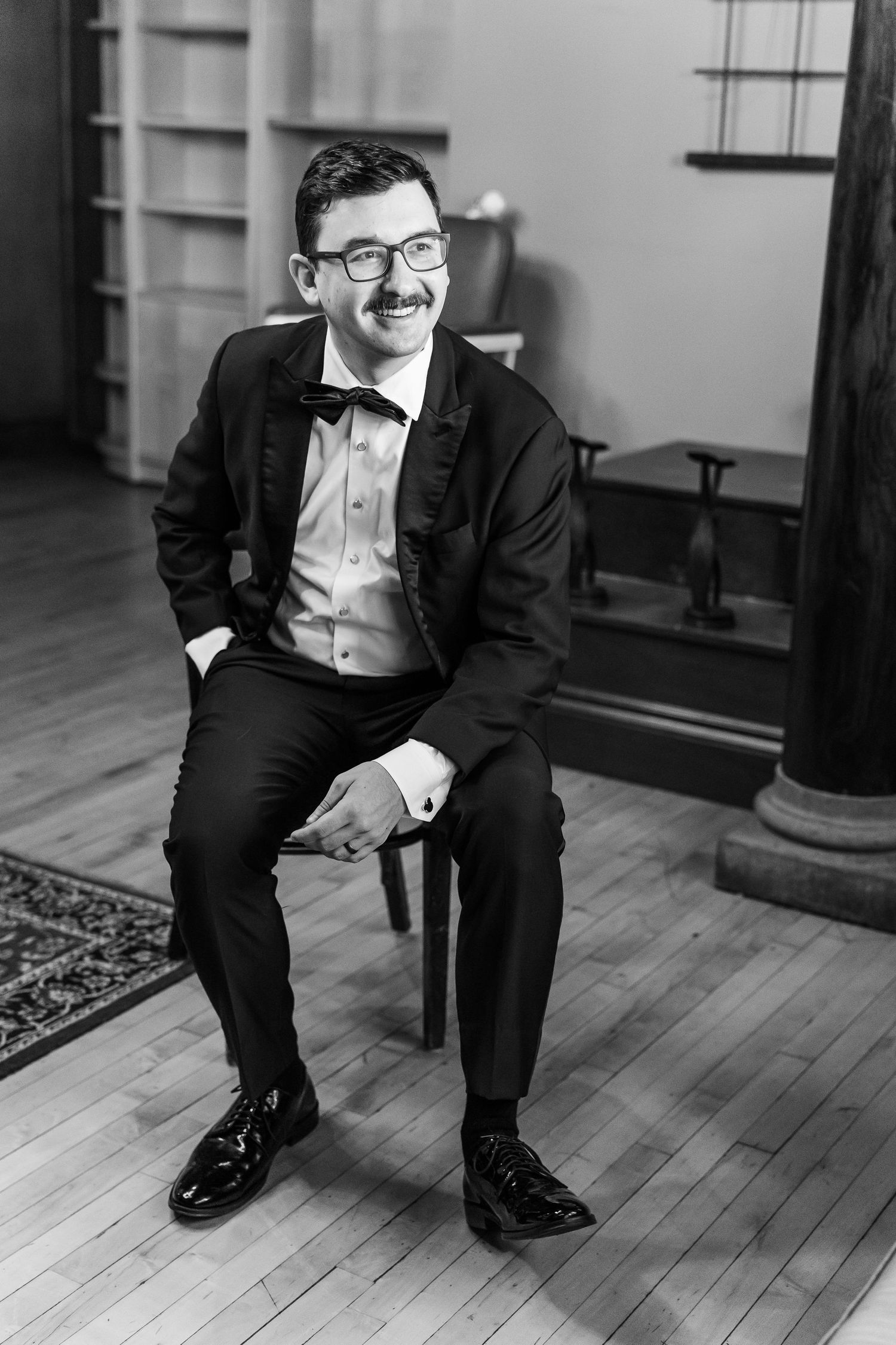 A person in a tuxedo sits casually on a chair, smiling in a formal indoor setting.