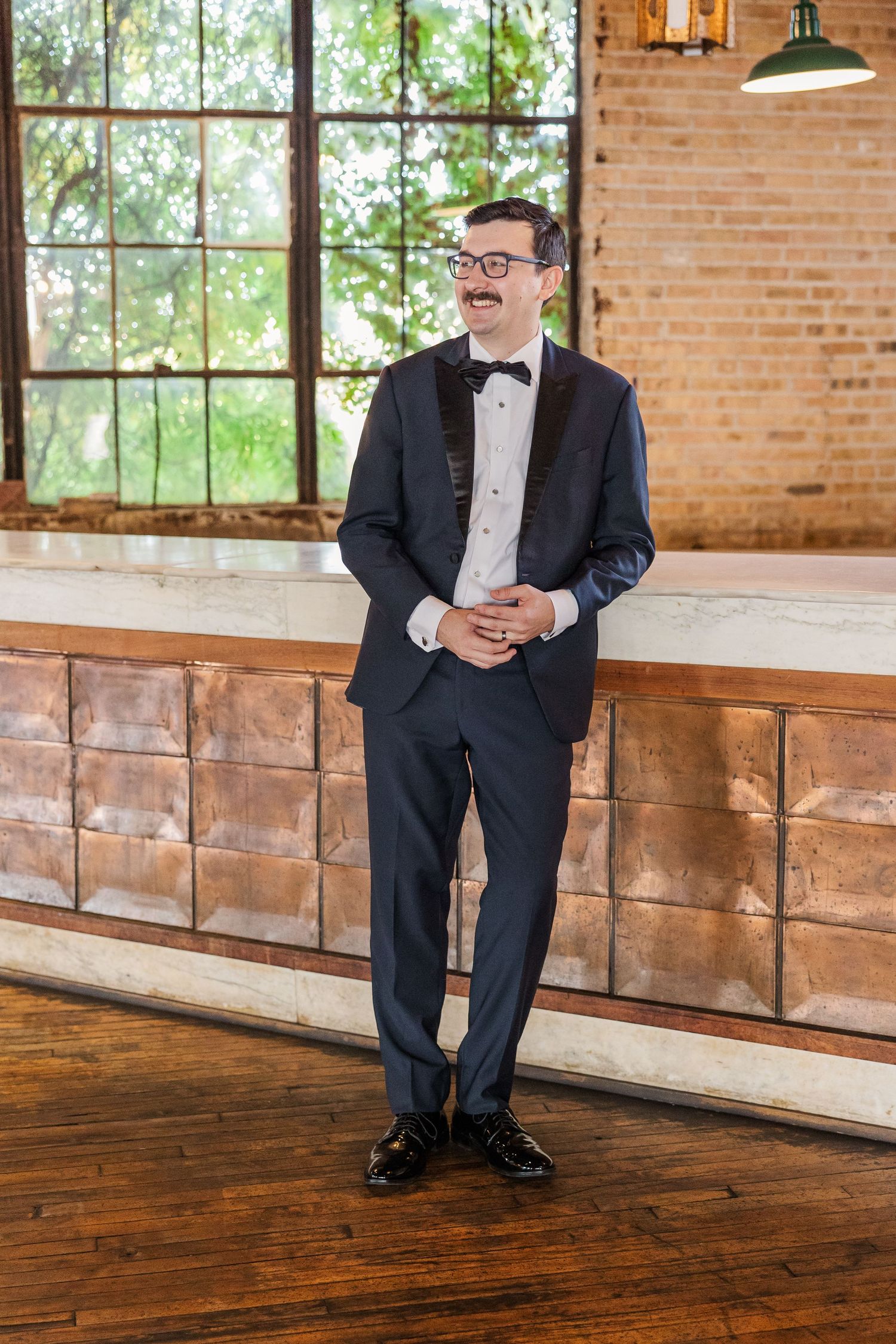 A person in formal attire stands against a rustic venue backdrop with large windows and exposed brick.
