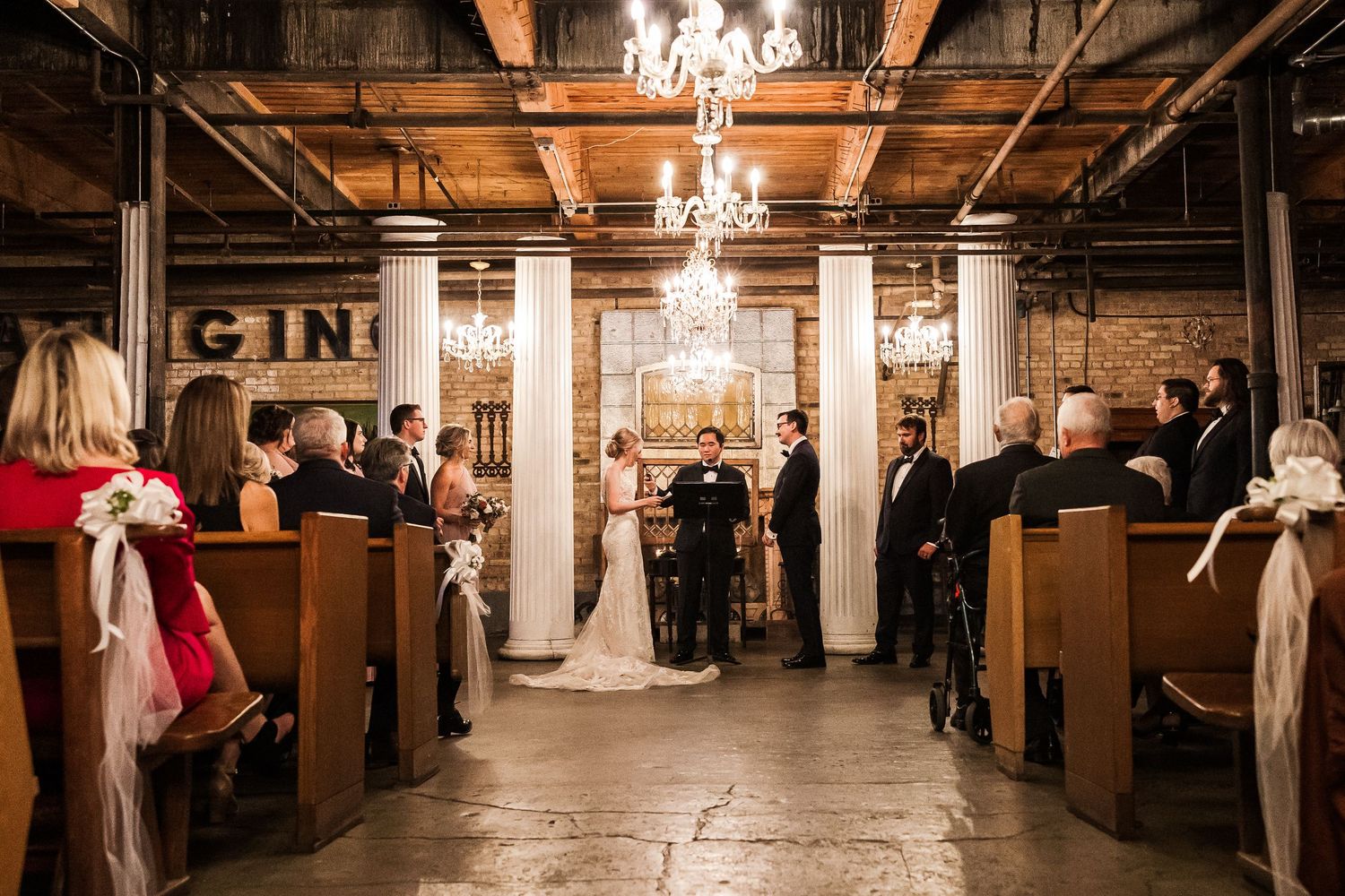 Wide view of wedding ceremony in industrial space with wooden pews and crystal chandeliers.