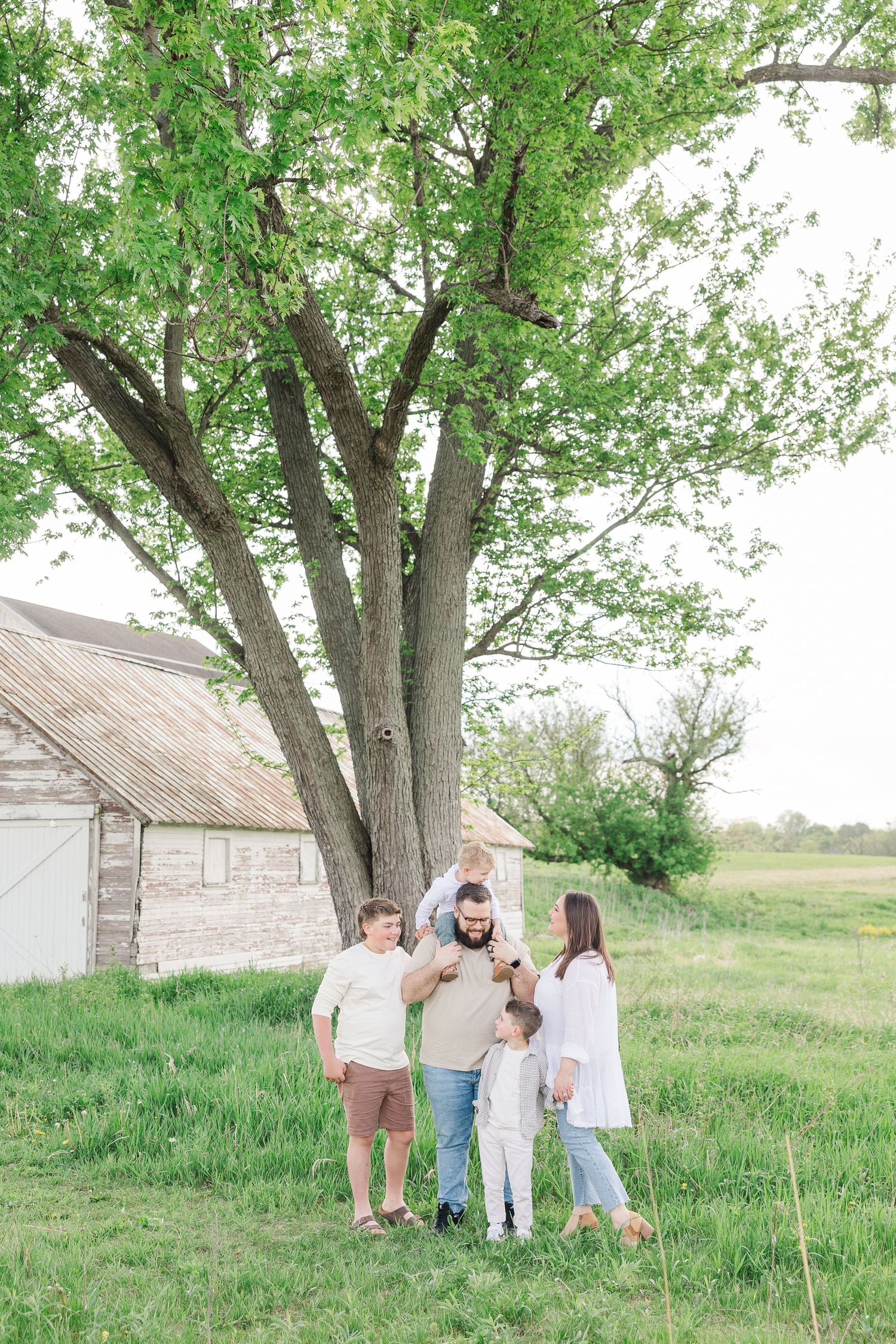 A family gathers beneath a large tree next to a rustic white barn in a pastoral countryside setting.