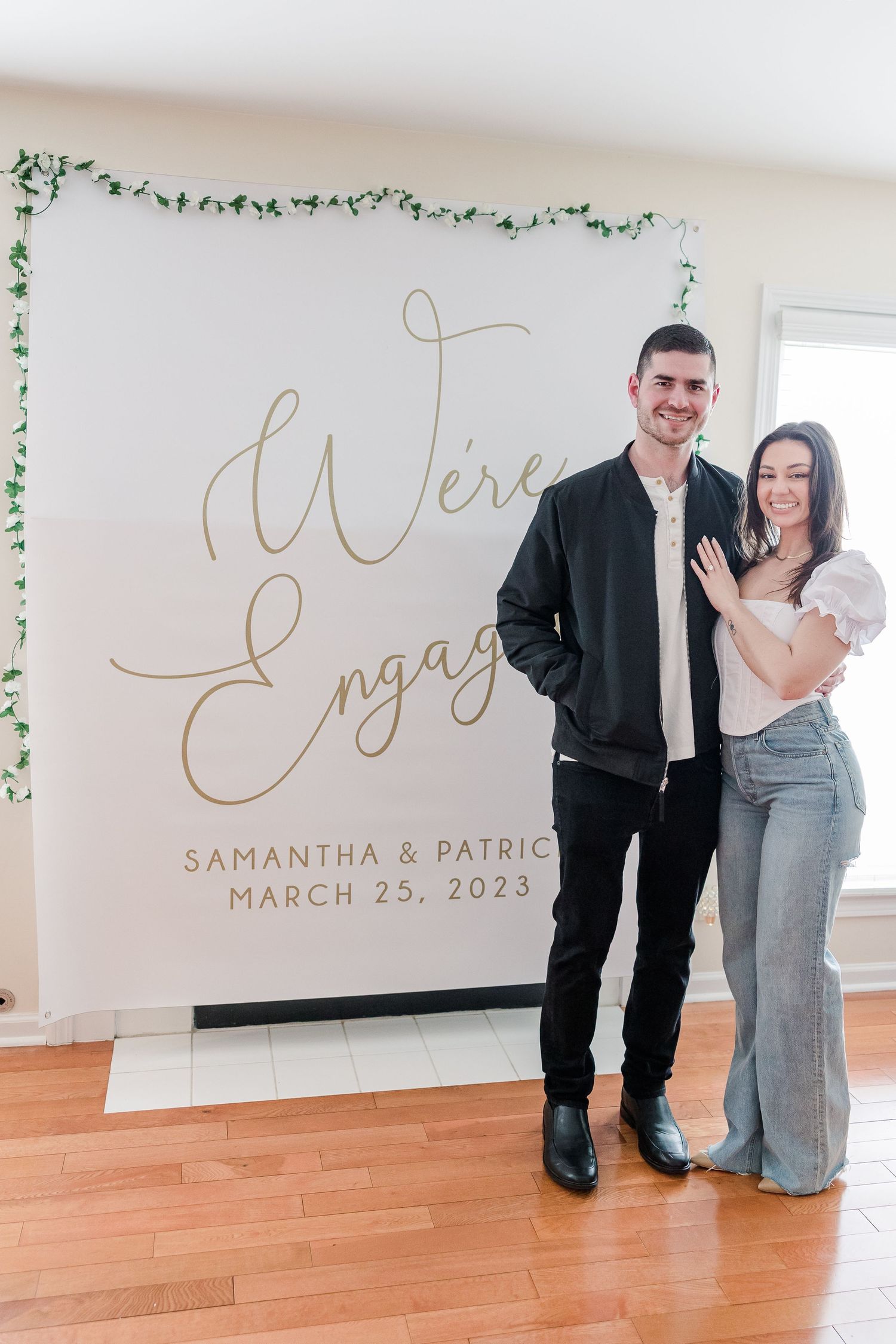 Couple standing together against elegant engagement announcement backdrop with decorative vines.