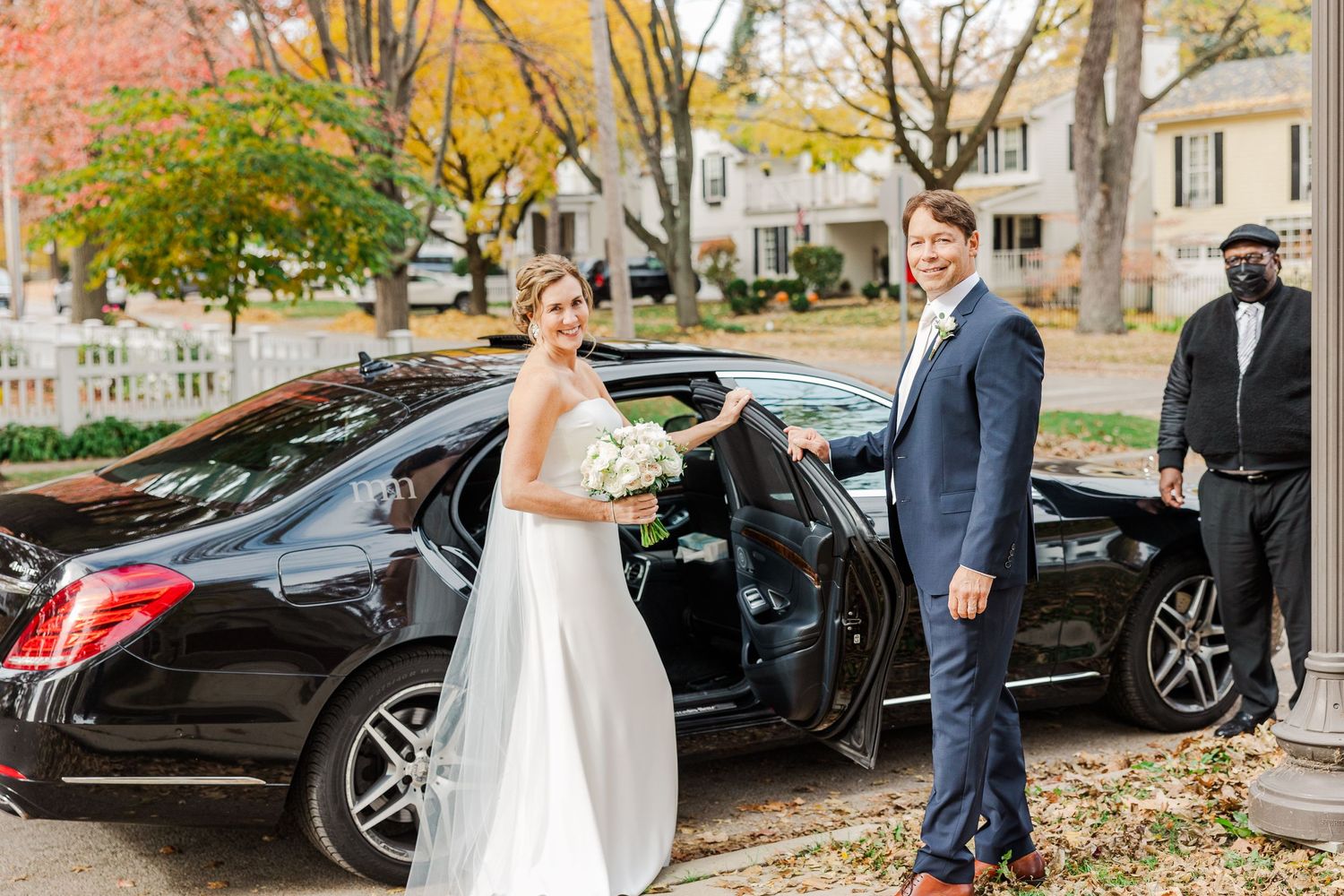 A newly married couple exits a black luxury car outside in autumn with colorful fall foliage in the background.