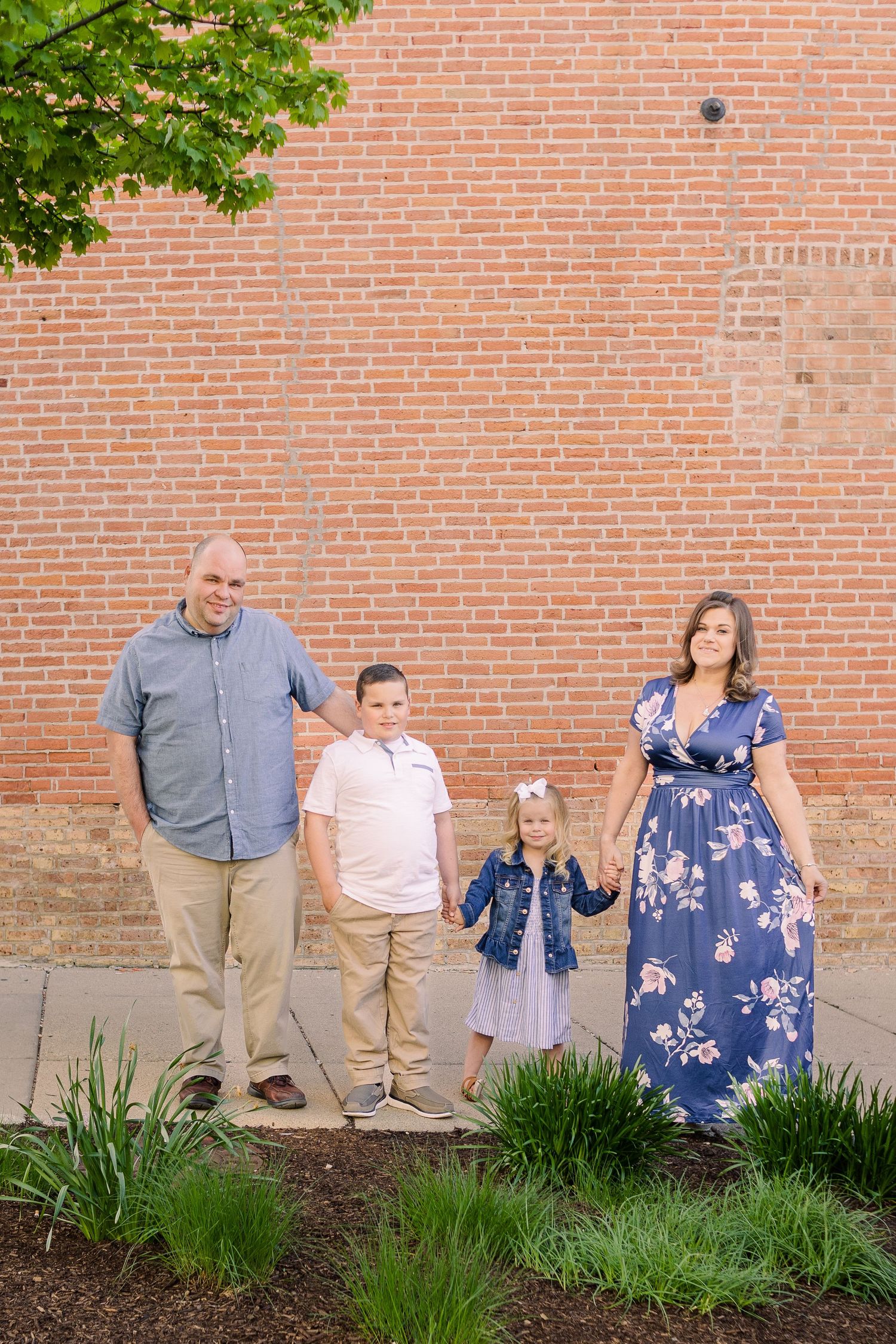 A family of four poses together against a brick wall with green plants in the foreground.