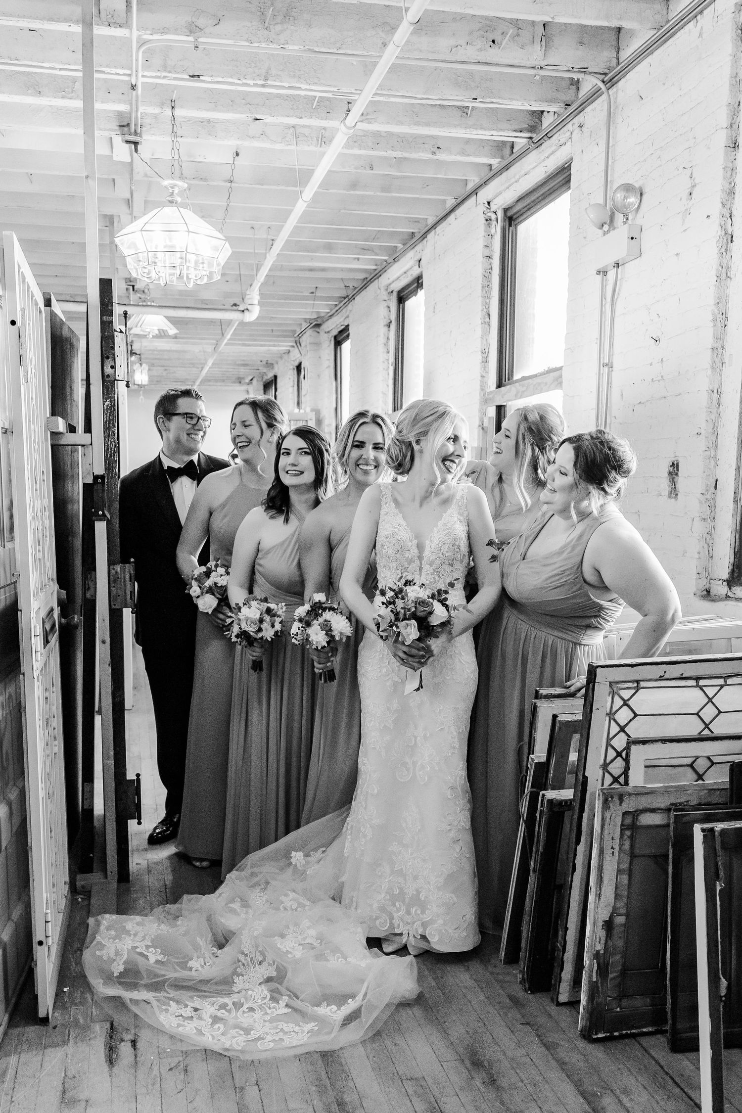 Black and white photo of bridal party standing in vintage hallway with exposed beams and chandelier.