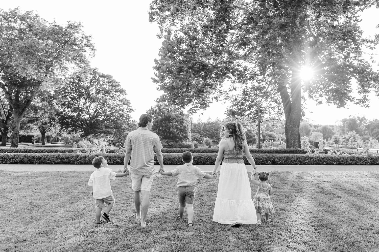 Black and white photo of family holding hands walking through lush park in warm sunlight.