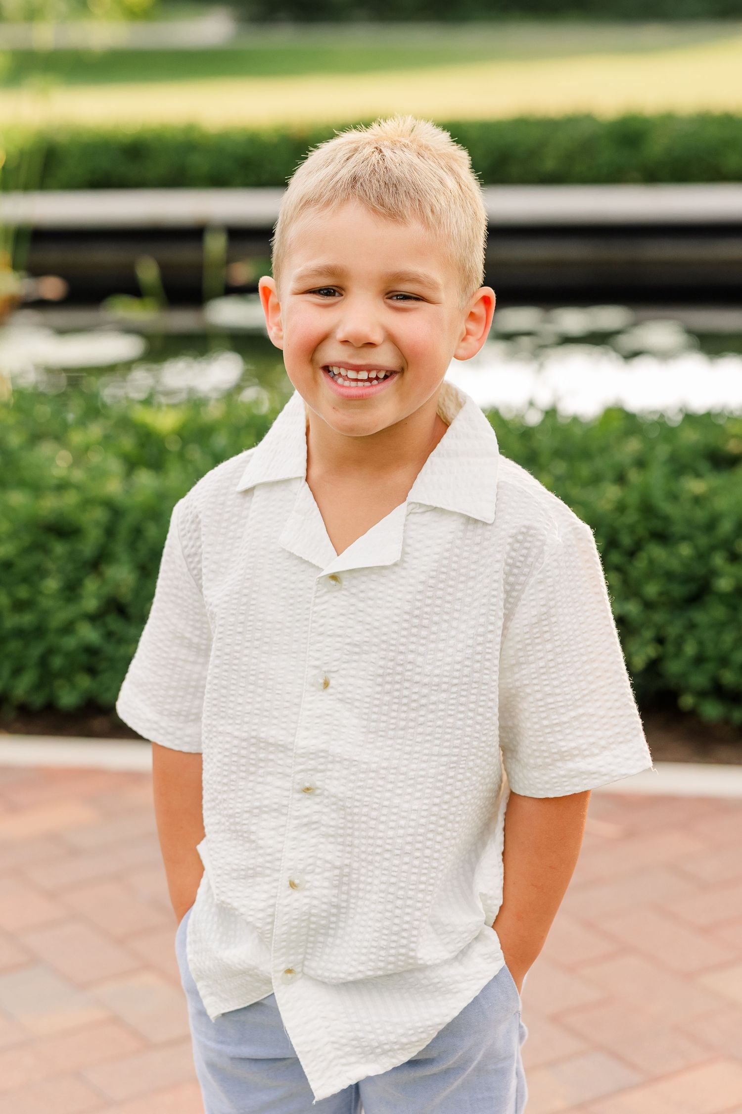 Smiling blonde boy in white textured button-up shirt standing outdoors in a garden setting.