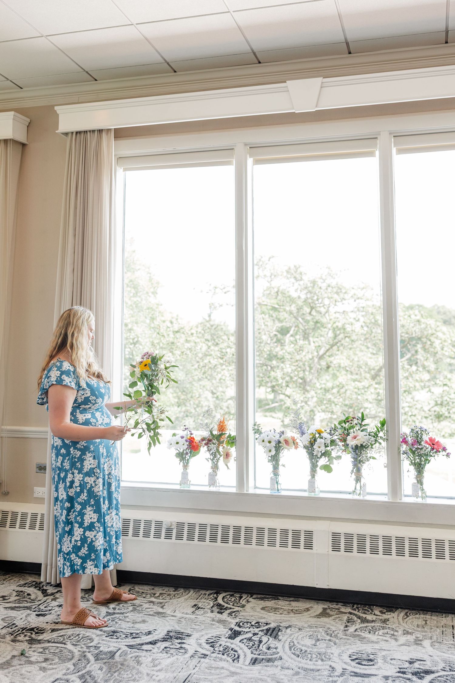 Woman in blue floral dress arranging colorful flower bouquets in vases along a large sunny window.