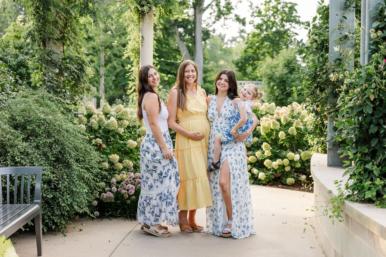 Three smiling women in floral and yellow dresses standing together in a lush garden setting.