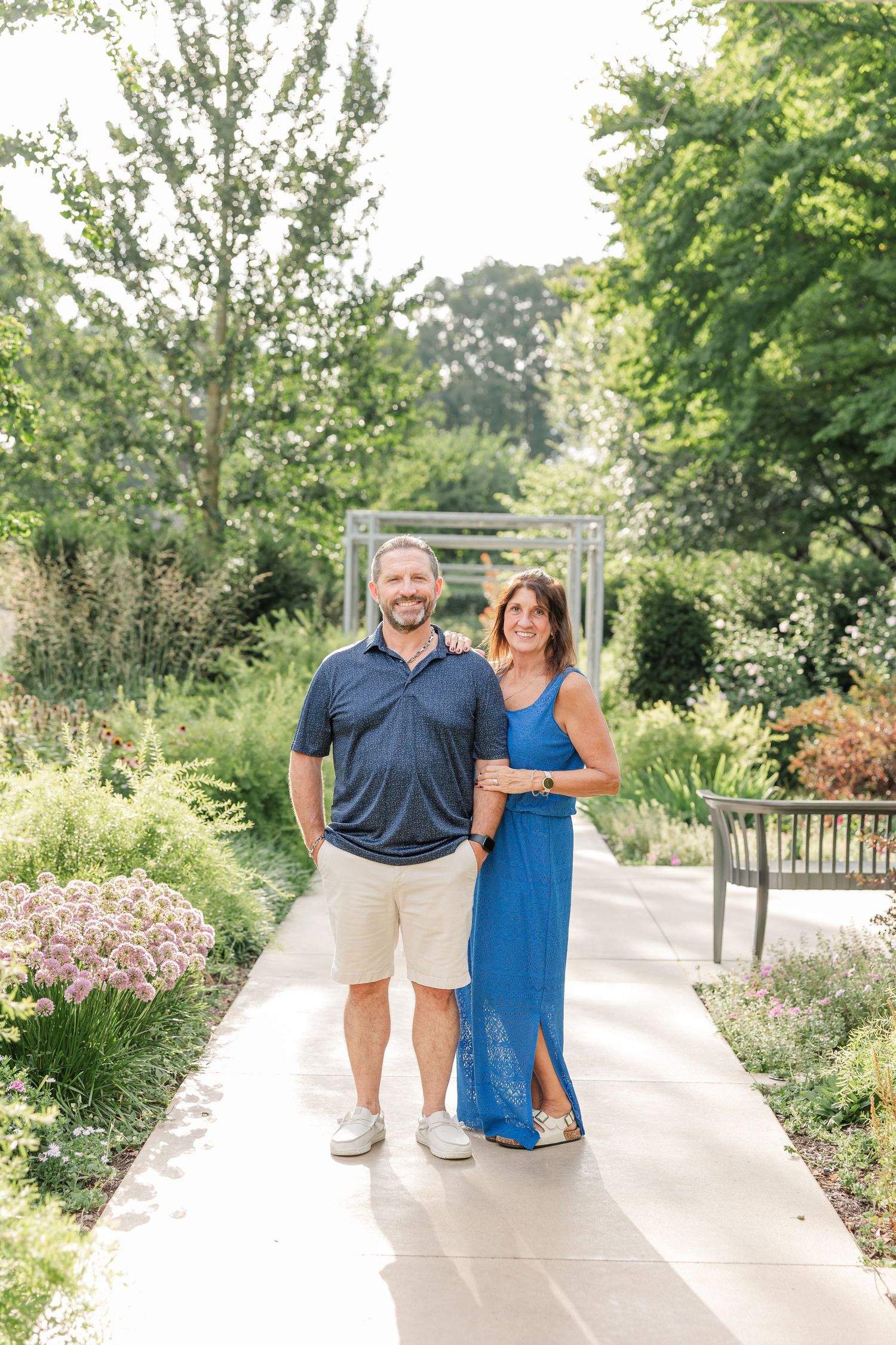 Happy couple posing on a sunny garden walkway surrounded by lush greenery and flowering plants.