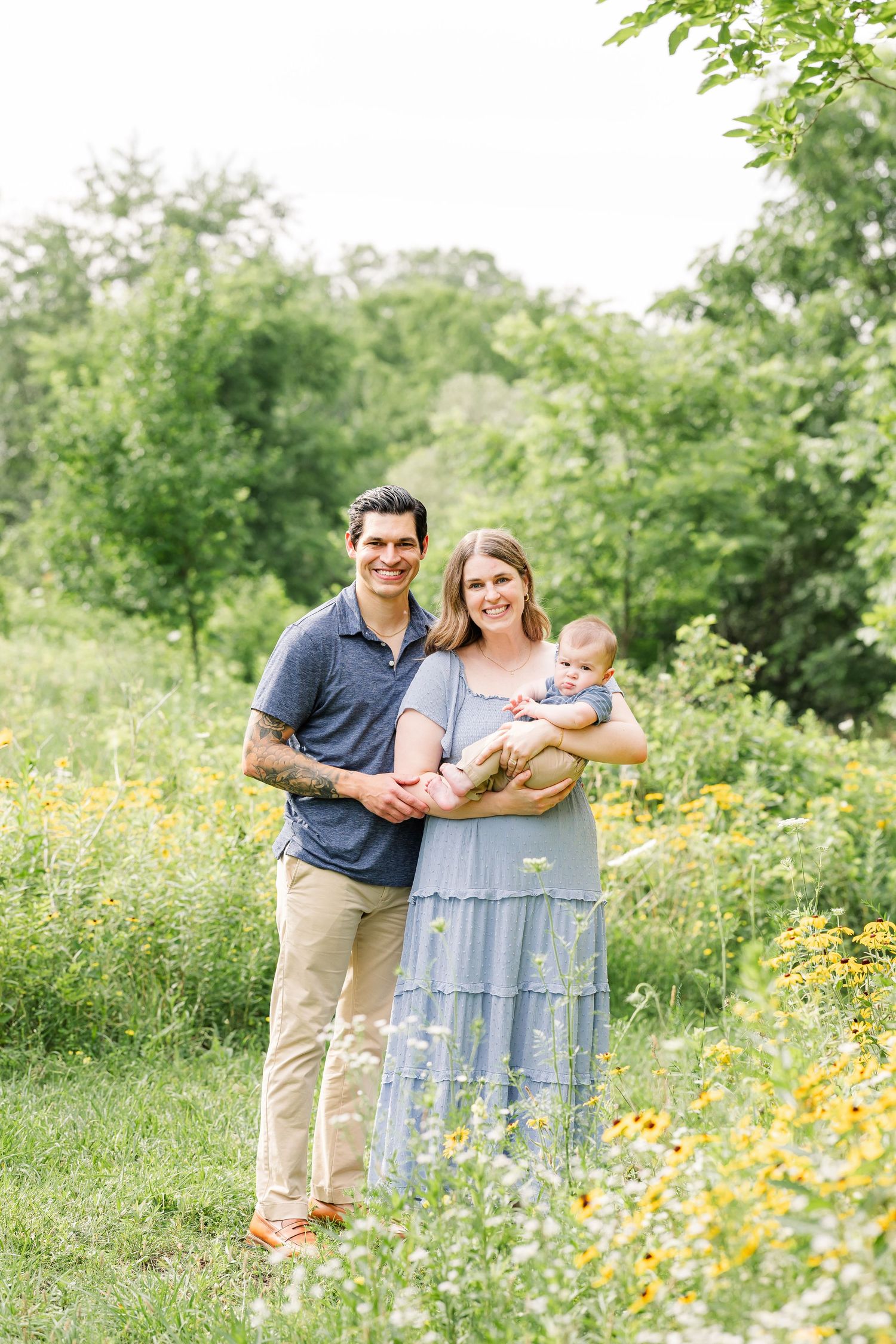 A smiling couple stands together in a wildflower meadow, the woman holding their newborn baby.