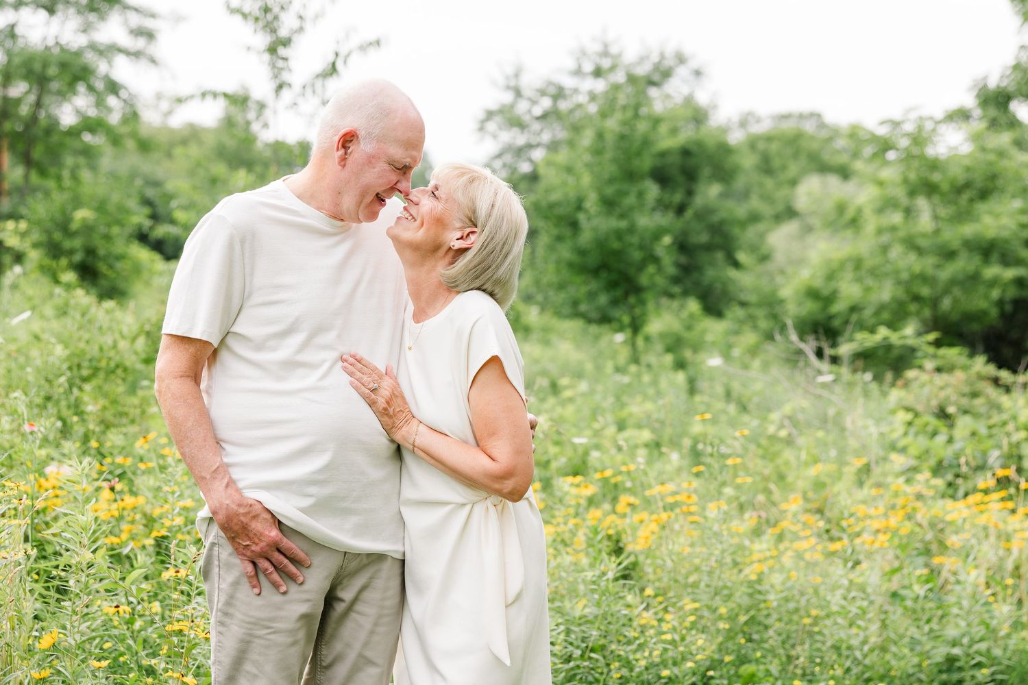 Elderly couple sharing a romantic moment in a sunny wildflower meadow, smiling at each other in white outfits.