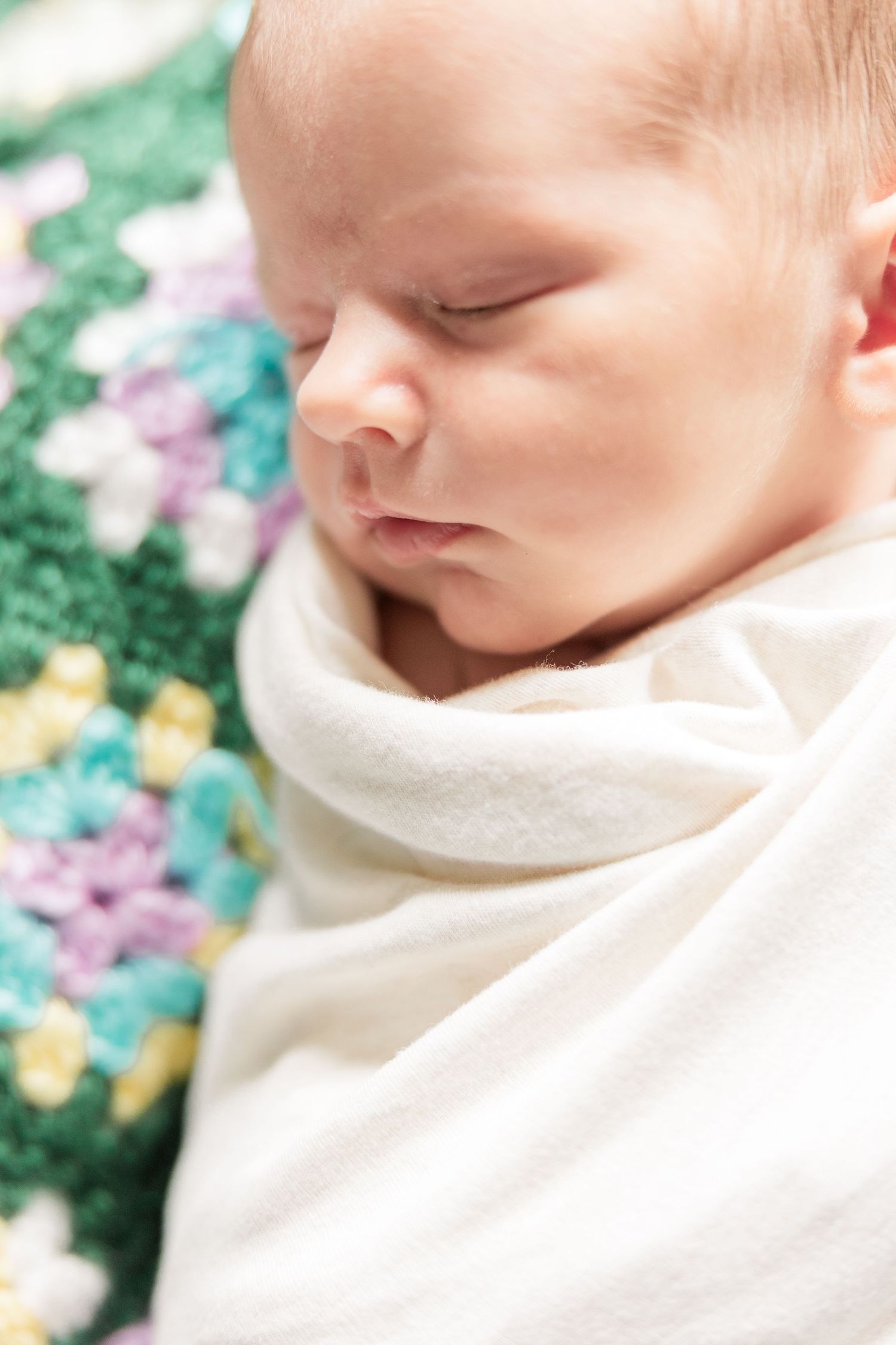 Close-up of peaceful sleeping newborn baby swaddled in white on colorful crocheted blanket.