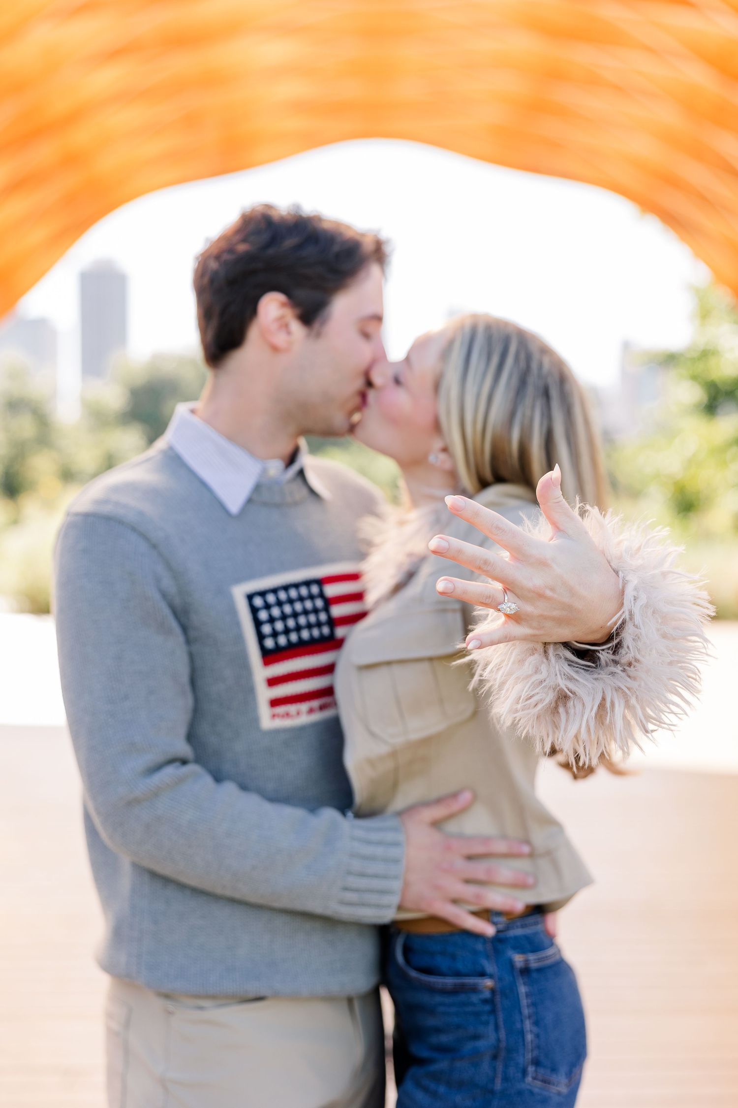 Couple sharing a kiss while showing off engagement ring under orange arch in Chicago's Millennium Park.