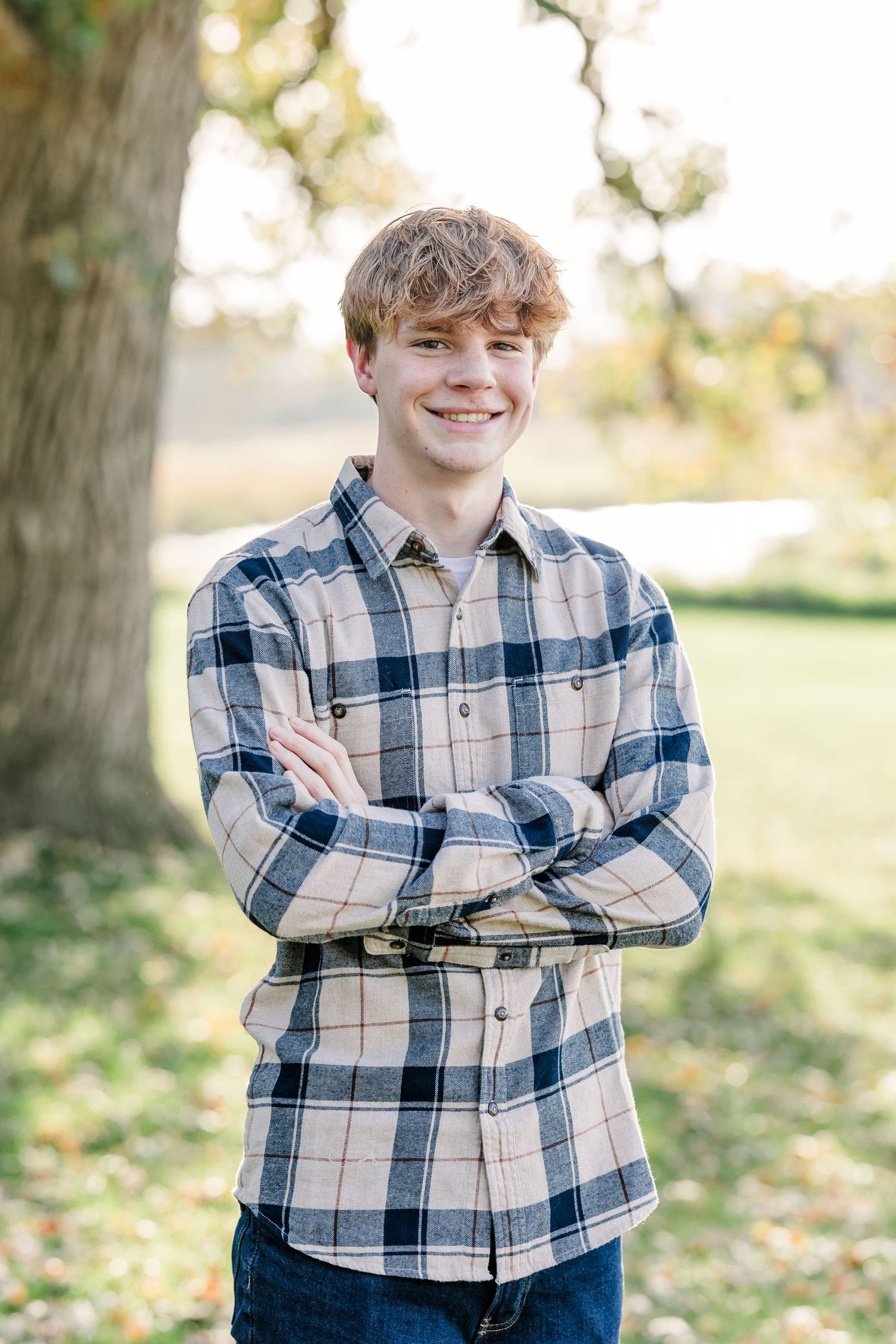 Smiling teenage boy with tousled hair stands with arms crossed in a plaid flannel shirt outdoors beneath a large tree in a sunny park.