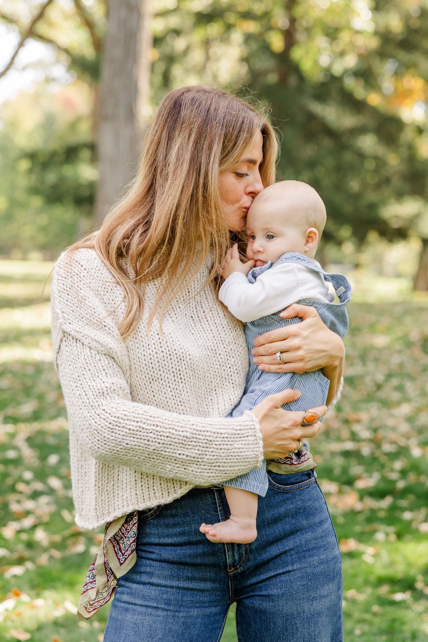 A mother in a cream sweater and jeans kisses her baby boy outdoors in a lush green park during autumn.