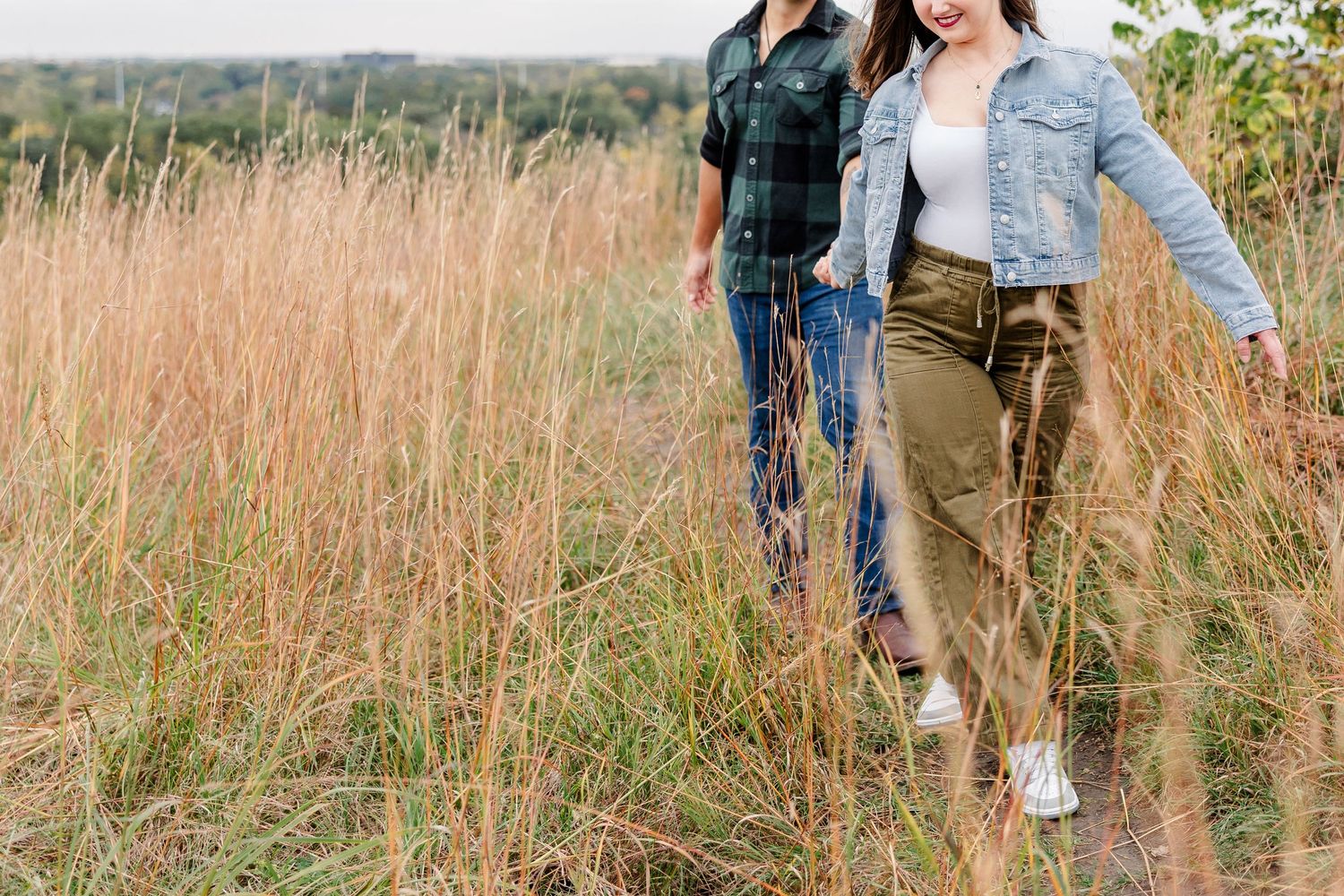 A couple in casual denim and green plaid walks hand in hand through tall golden grass on an overcast autumn day.