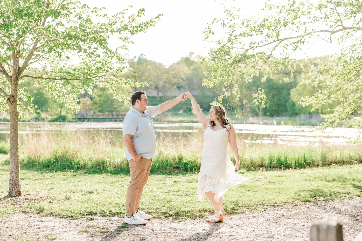 Couple dancing outdoors near a sunlit lake, man in gray shirt spinning woman in white dress among green trees.