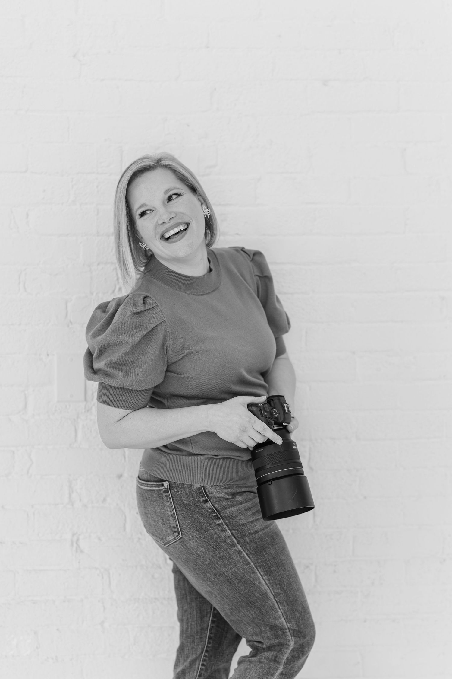 Black-and-white branding headshot of Cassie Lacina holding her camera at The Studio Chicago against a white brick wall