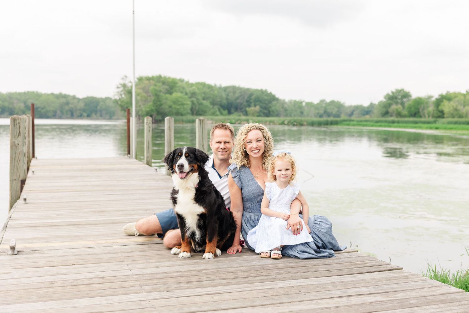 A family and their Bernese Mountain Dog sit together on a wooden dock by a serene lake on a summer day.
