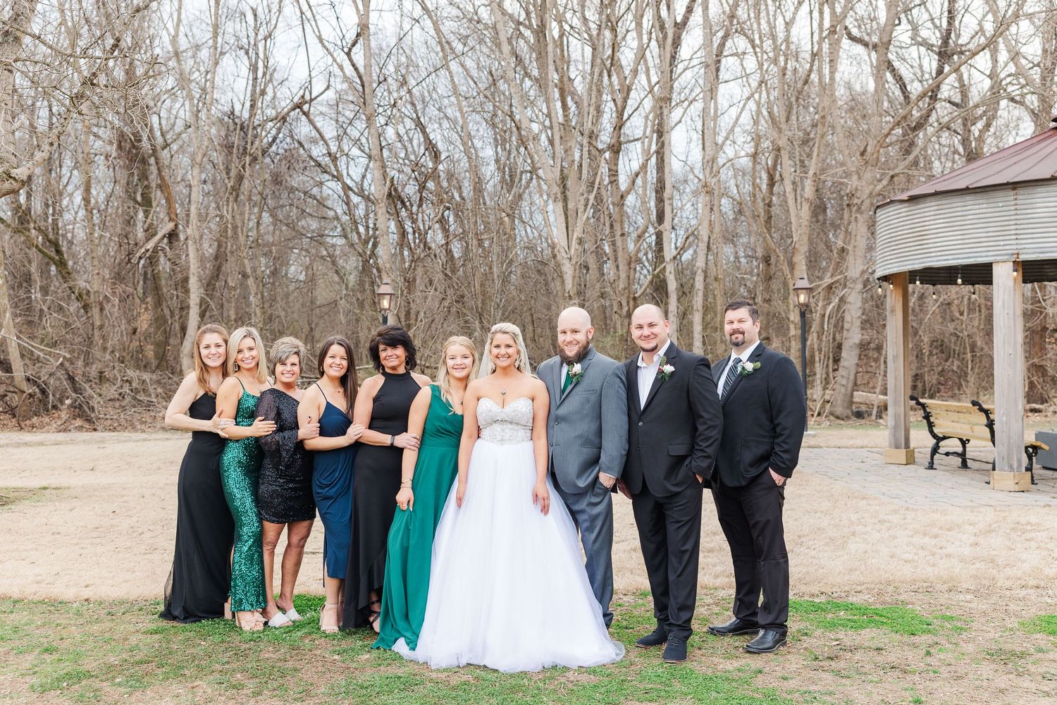 Wedding party poses outdoors in formal attire with bridesmaids in jewel-toned dresses and groomsmen in black suits.