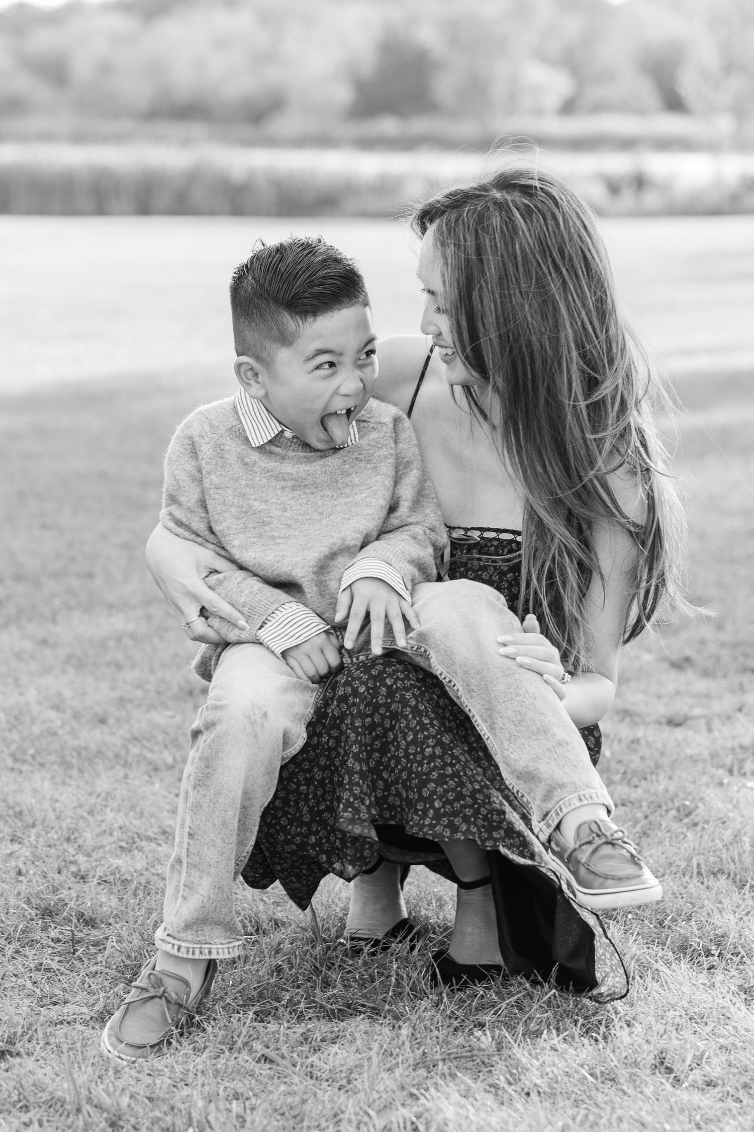 A couple sits together on grass in black and white, sharing a playful moment outdoors. Busse Woods