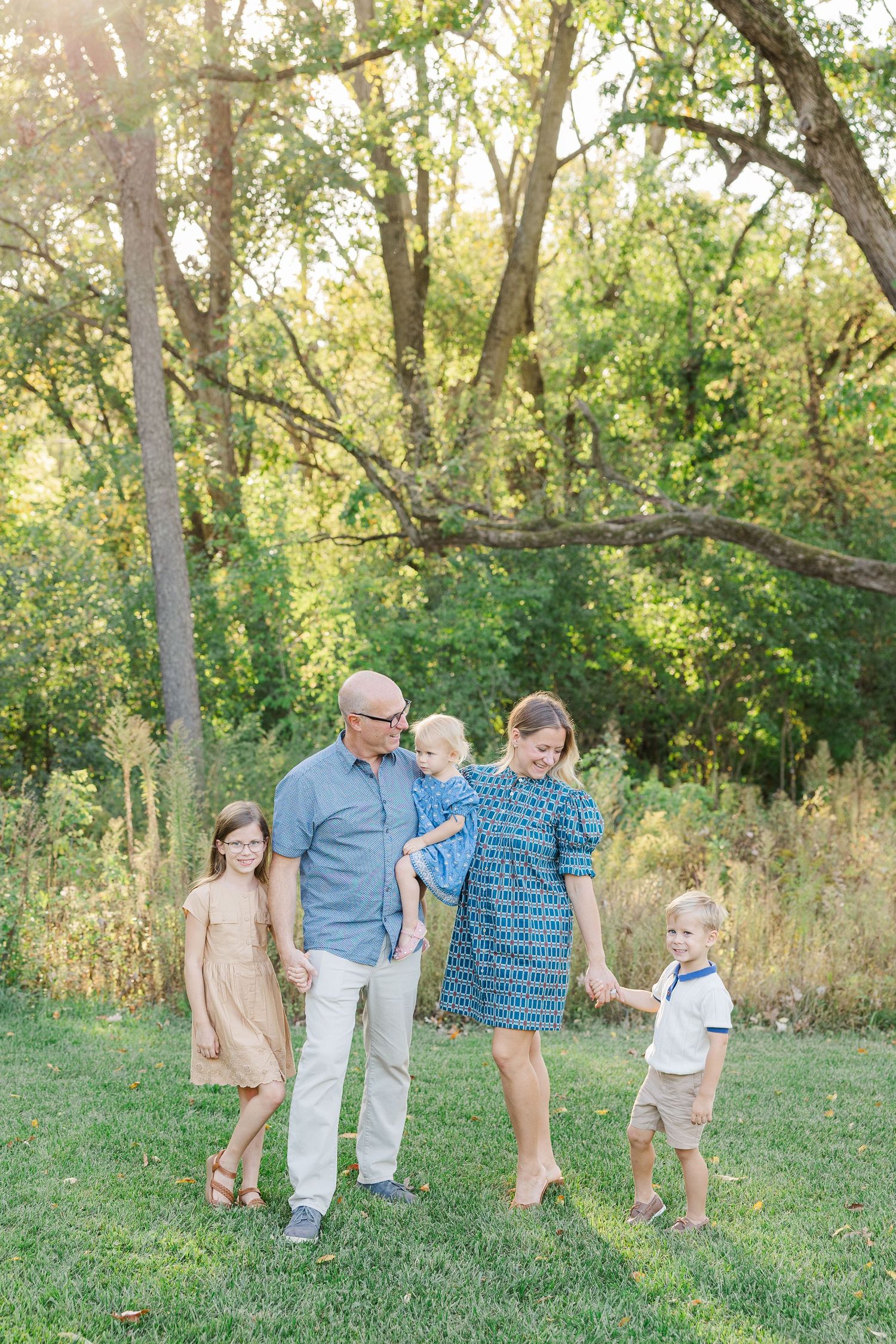 A family walking together through a sunlit wooded area wearing coordinated blue and beige outfits.