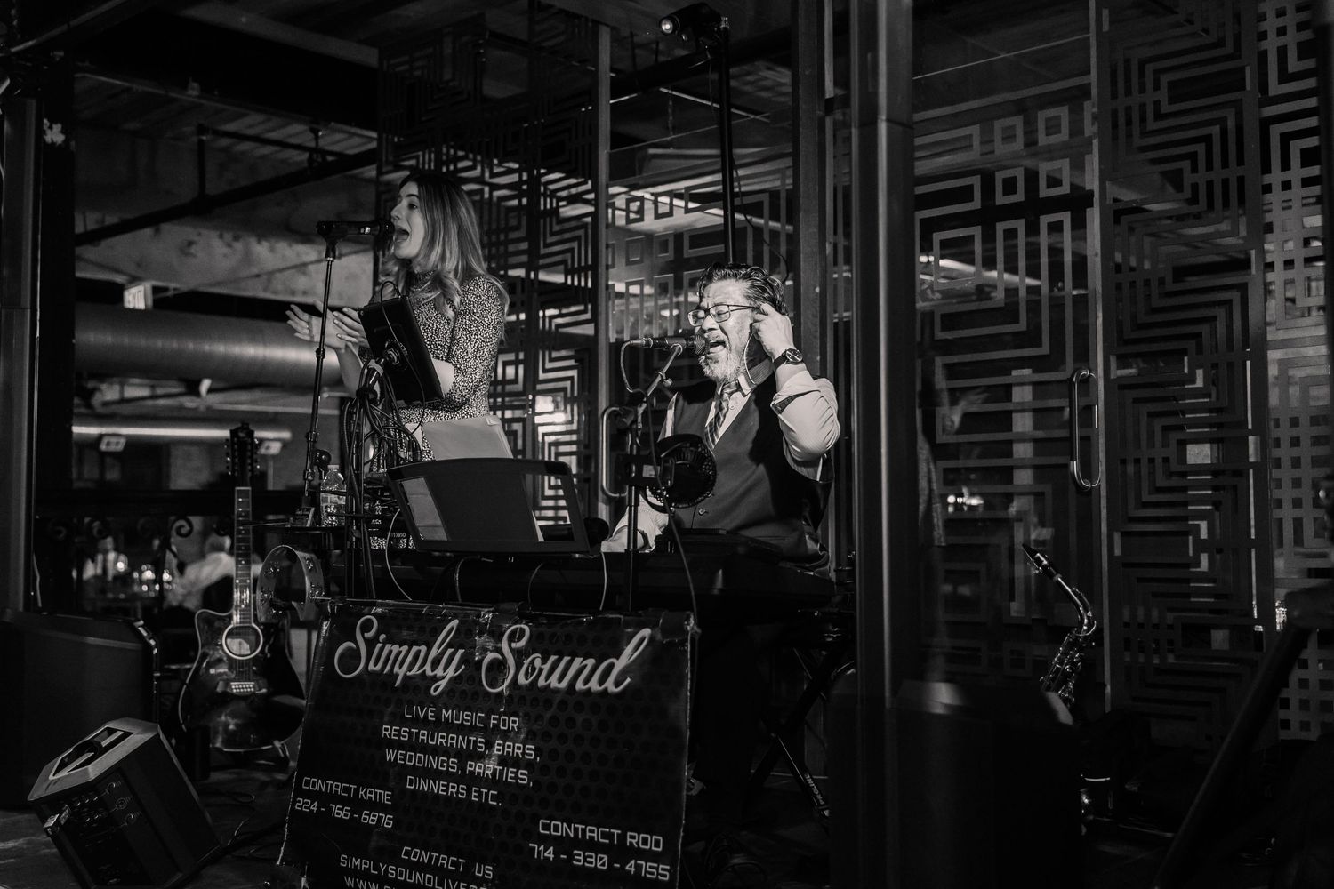 Black and white photo of musicians performing on stage with patterned wall backdrop.