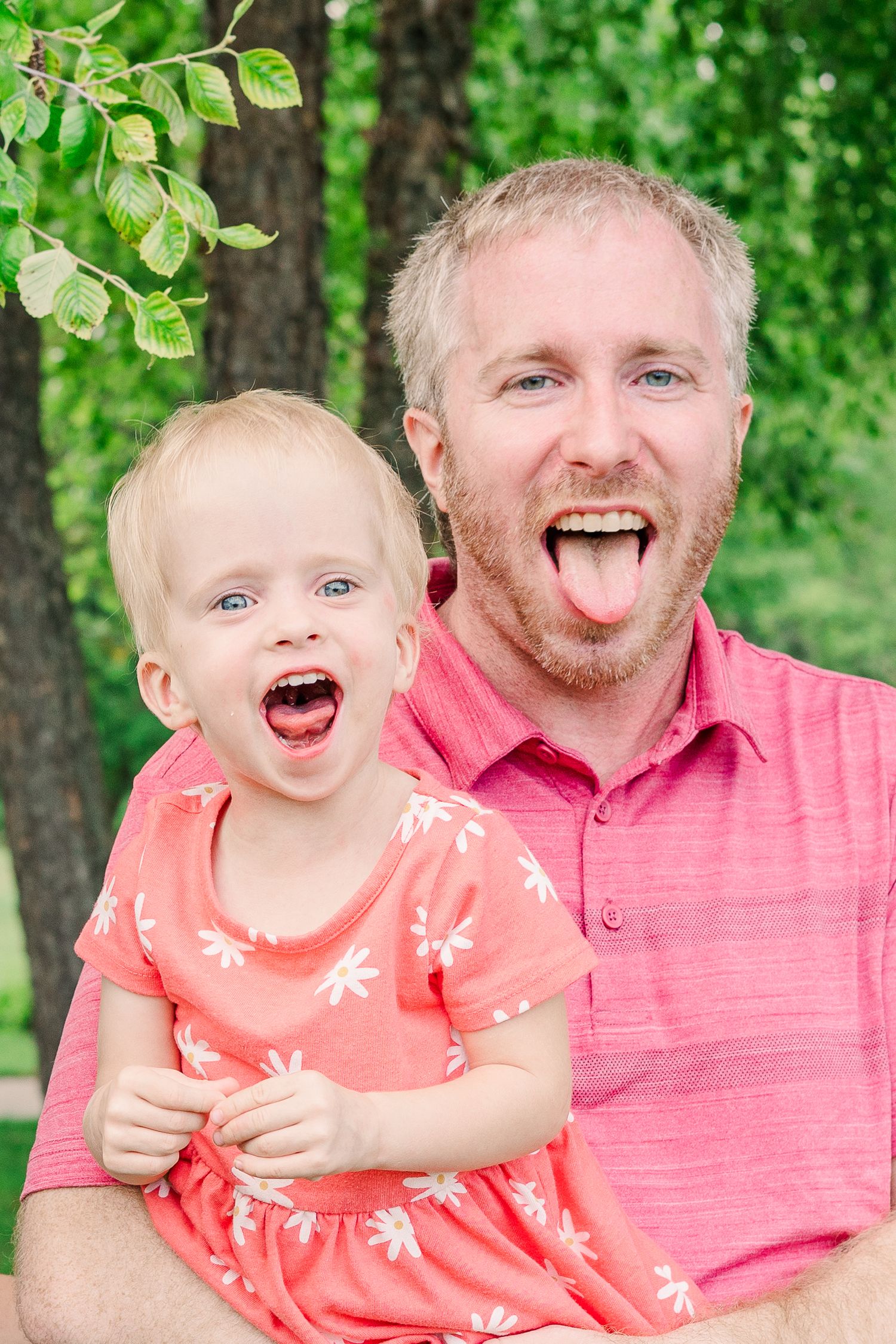 A parent and child stick out their tongues playfully while taking a photo outside.