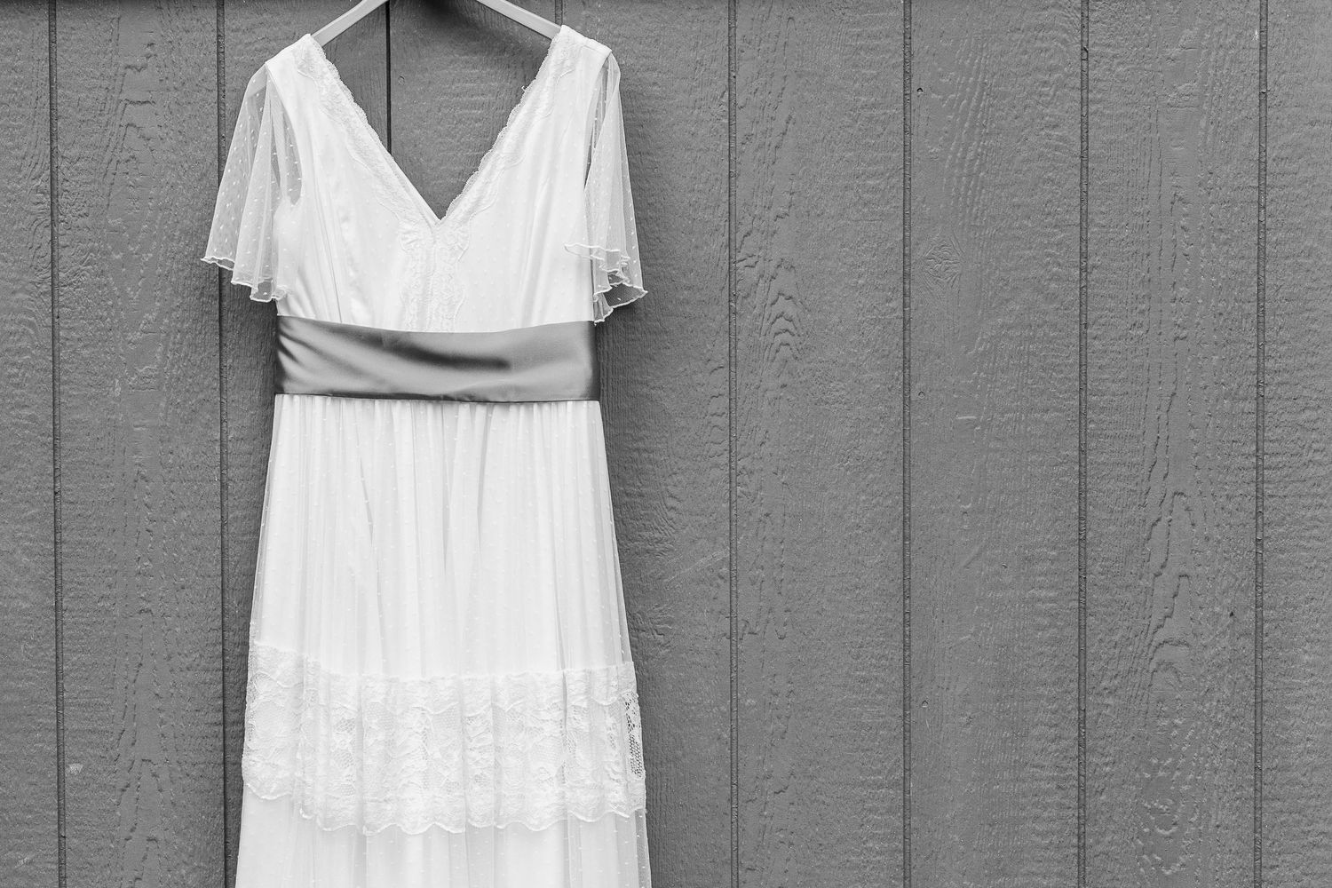 A vintage-style white wedding dress with flutter sleeves hanging against a grey wooden wall.