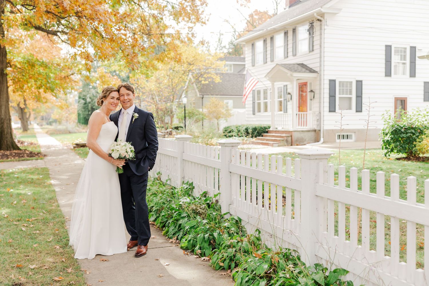 Wedding couple poses beside white picket fence during fall with classic house in background.