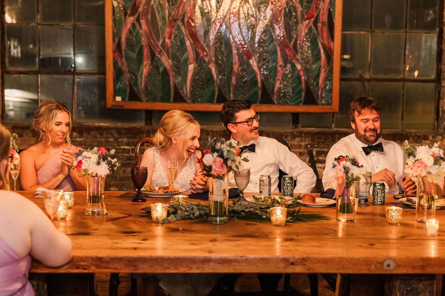 Wedding guests laugh and celebrate together at a rustic wooden table during reception dinner.