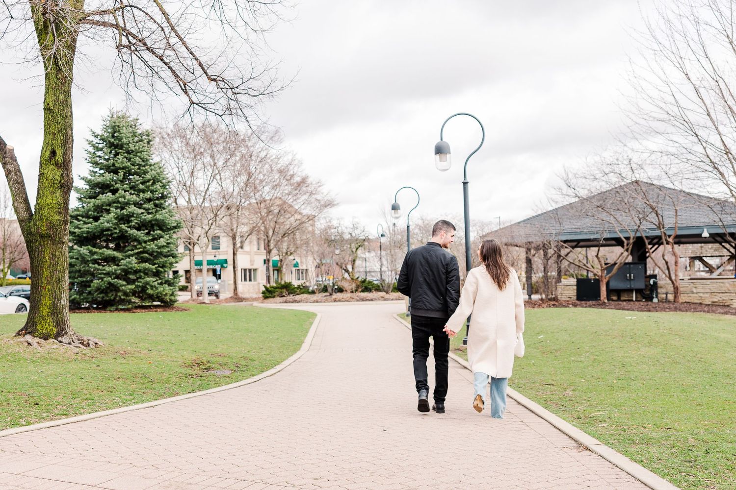 A couple walks along a winding brick path through a park with evergreen trees and lamp posts.