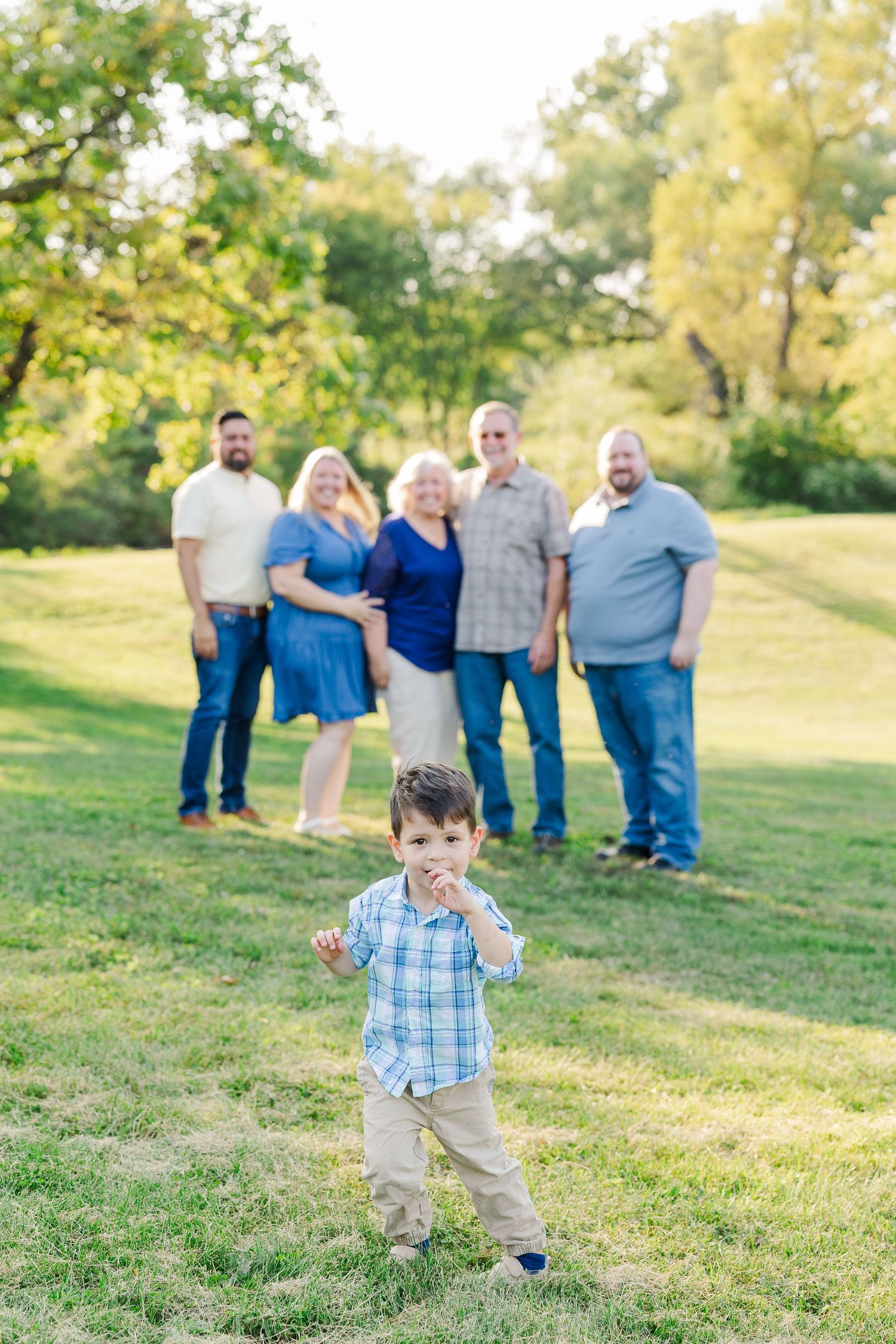 Group of adults gathered on grassy hillside while young child in plaid runs and plays in foreground.Sorg family portraits at Shamrock Hills
